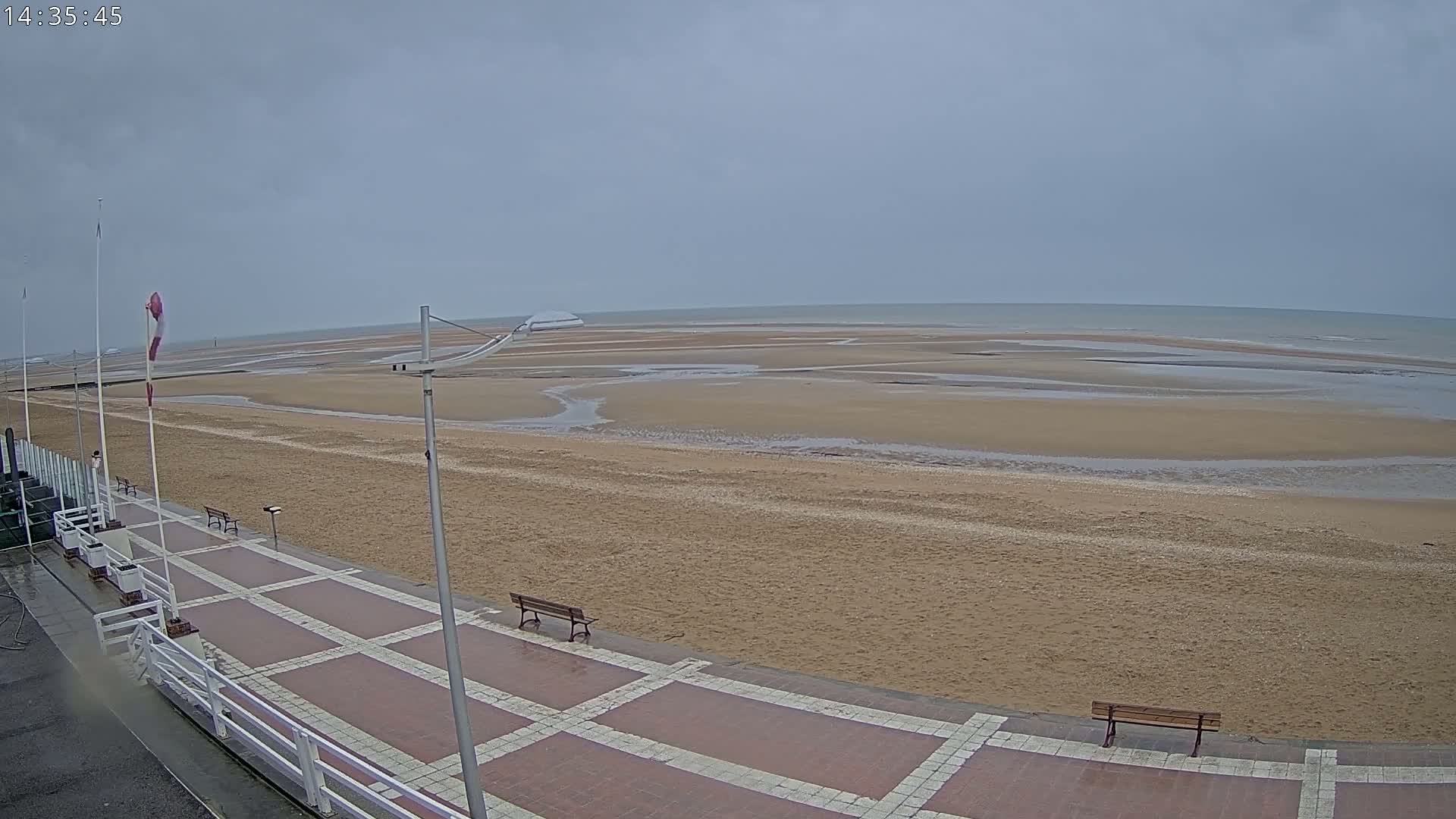 A wide sandy beach stretches to a calm, gray sea under an overcast sky, with a paved promenade featuring benches visible in the foreground.