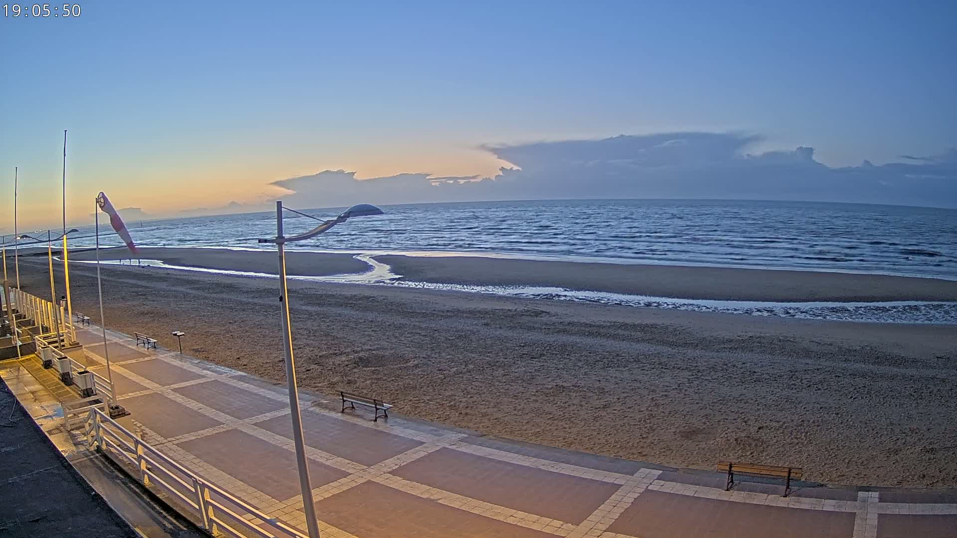 A sandy beach with a paved walkway and benches is visible at dusk under a mostly clear sky with some clouds on the horizon, overlooking a slightly wavy ocean.