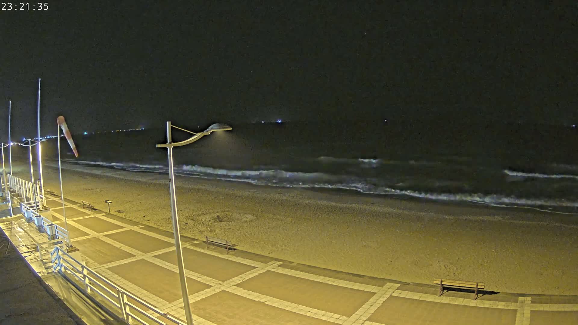A nighttime view of a sandy beach with gently rolling waves under a clear, dark sky, illuminated by lights along a paved walkway.