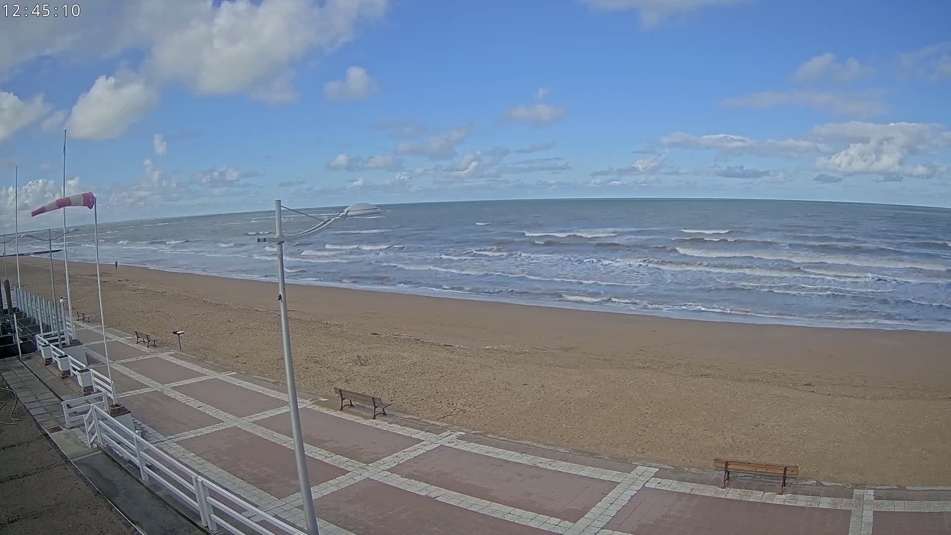 A sandy beach with a paved walkway and benches is seen under a partly cloudy sky, with moderately sized waves rolling into the shore.