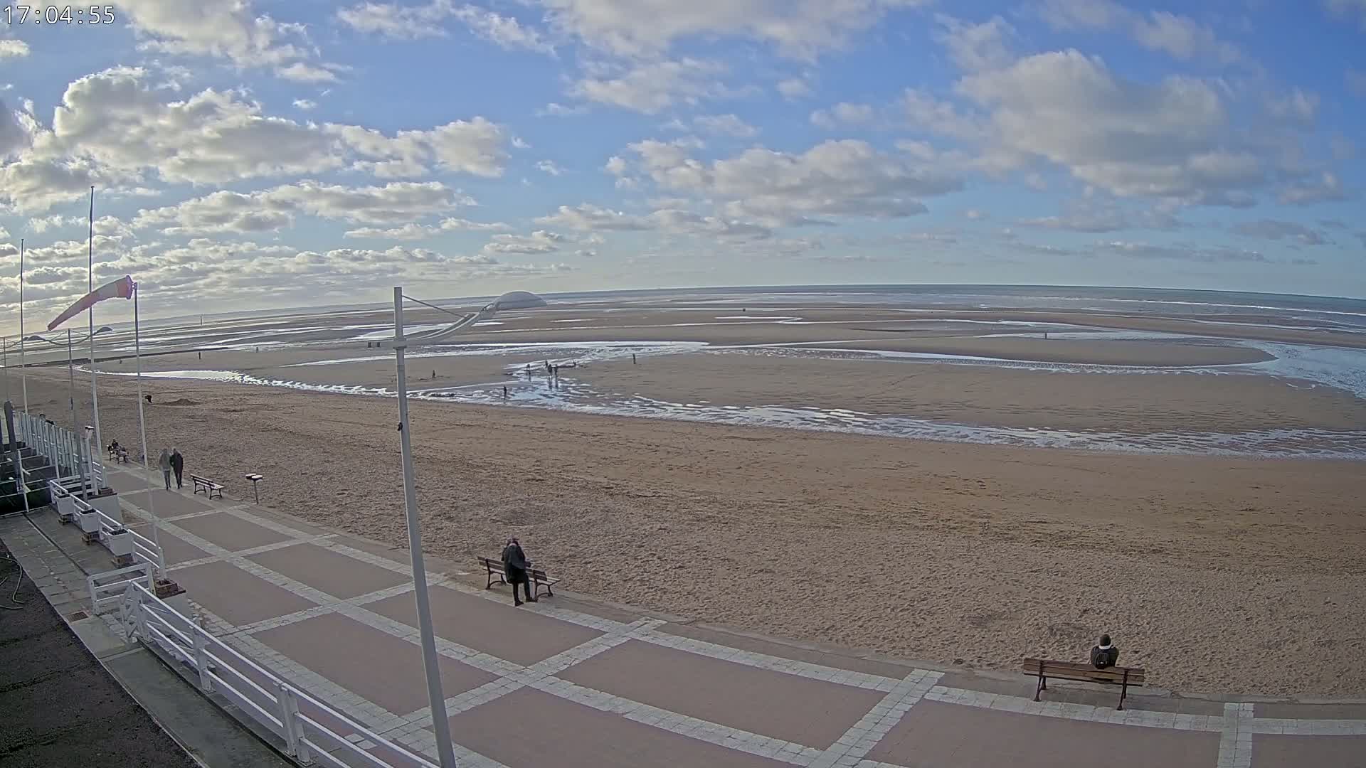 A mostly sunny day shows a wide sandy beach with a paved walkway, a few people, and a calm ocean receding to reveal tidal pools.