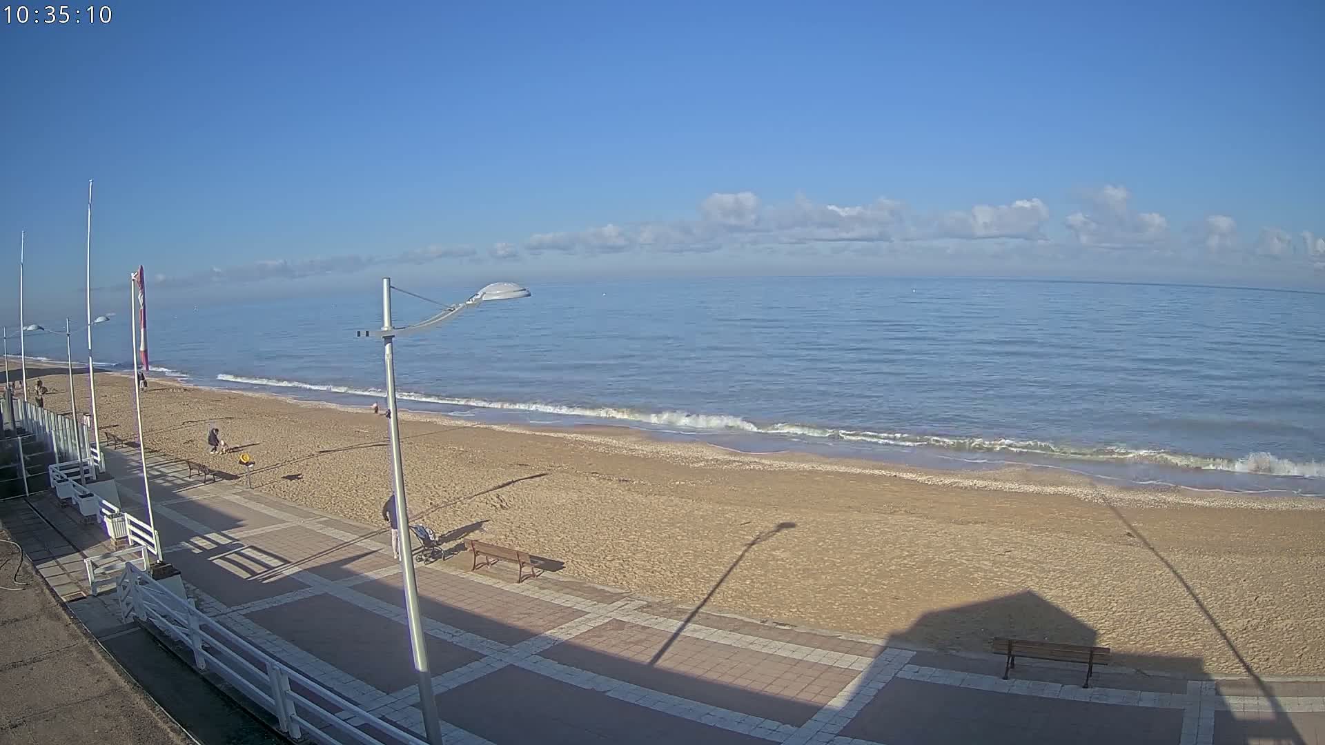 A sandy beach with a paved walkway and a few people is visible, under a mostly clear blue sky with some scattered clouds, and calm waves lapping the shore.