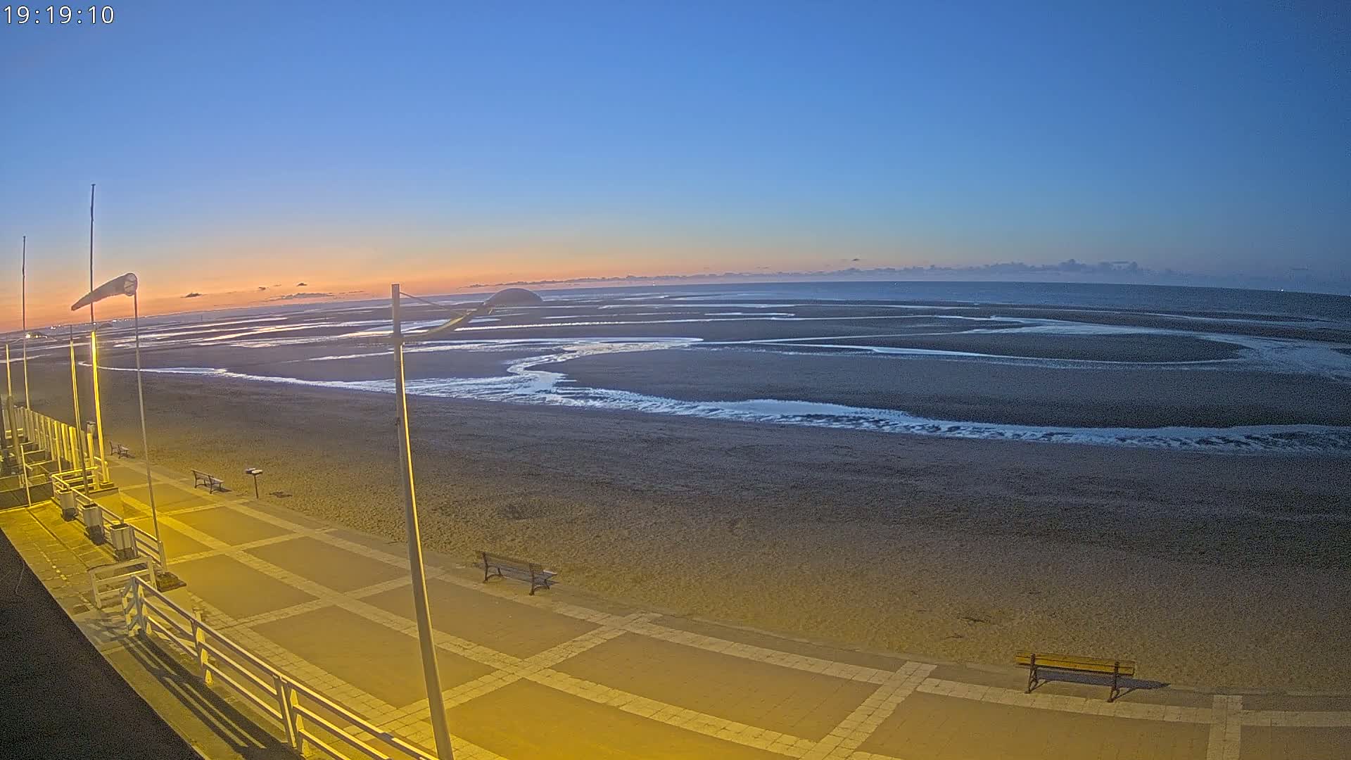A beach at dusk, with a paved walkway, benches, and a windsock, under a clear sky.
