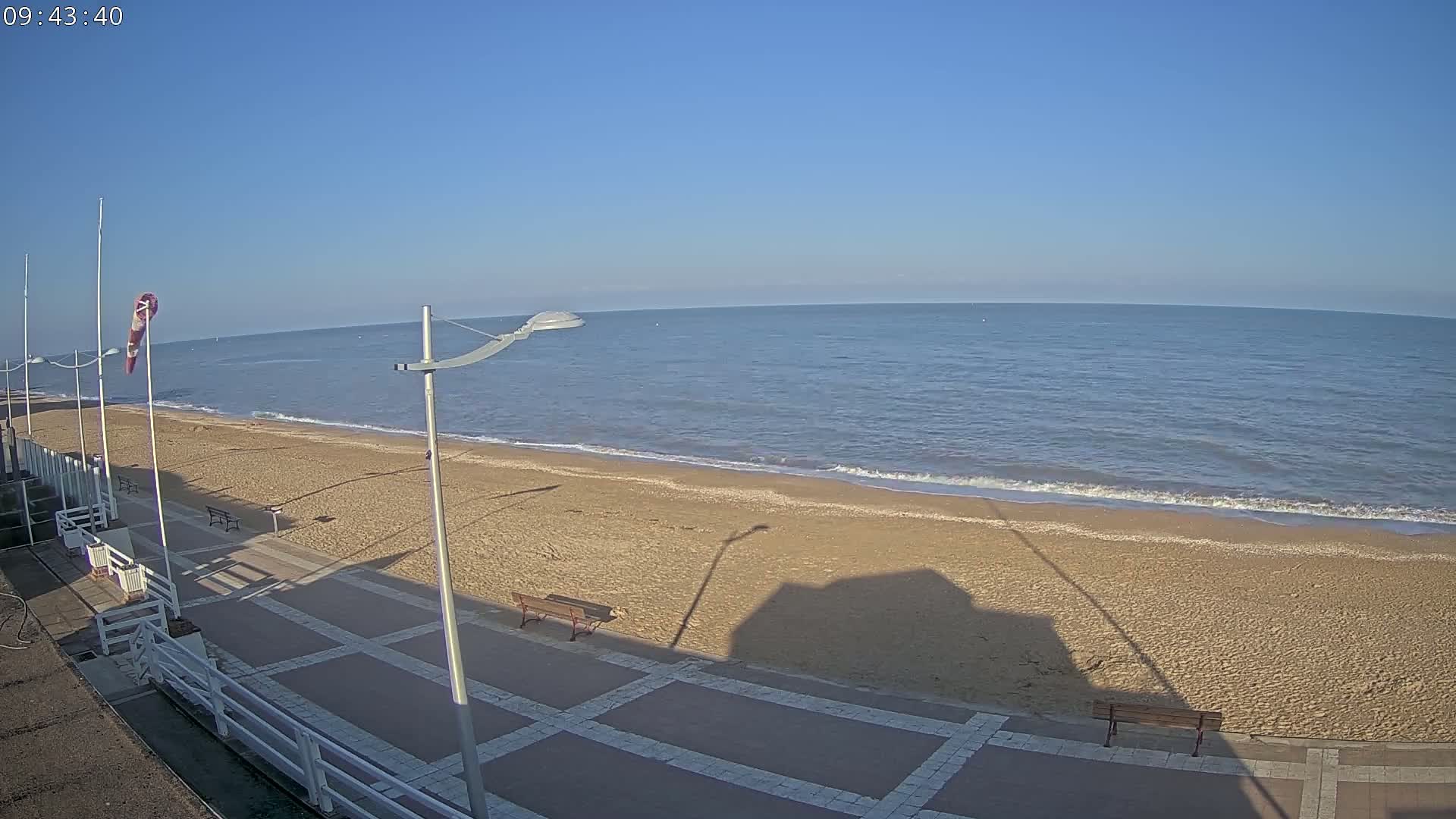 A sandy beach with a paved walkway and benches is shown, overlooking a calm ocean under a clear blue sky.