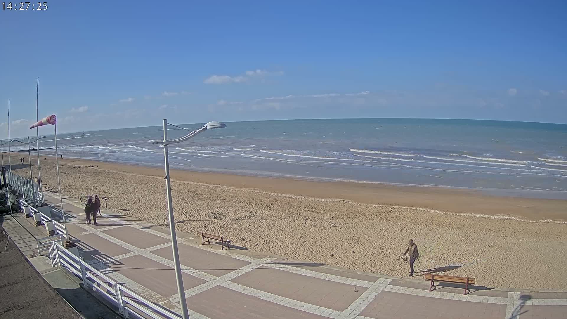 A sandy beach with a paved walkway and several people walking, under a mostly sunny sky with a moderately windy ocean.