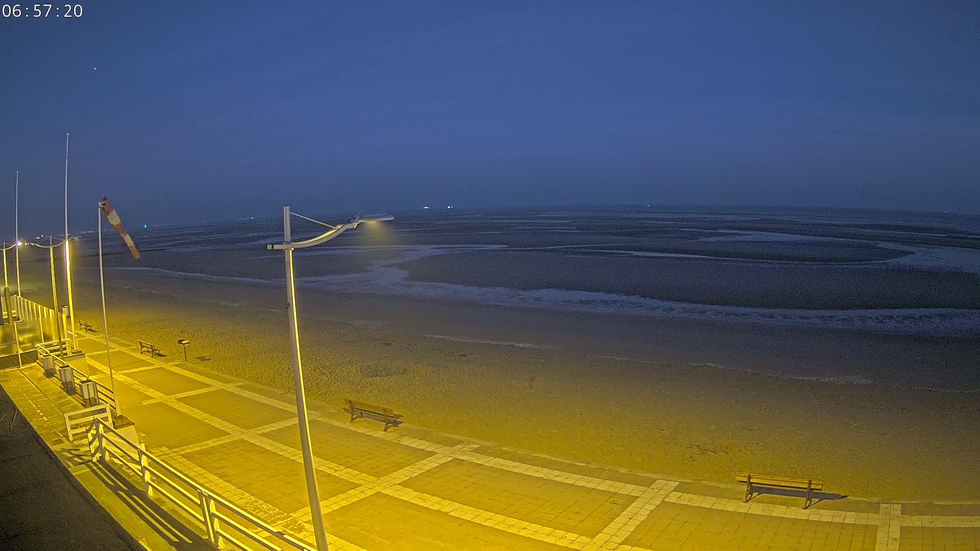A mostly empty, sandy beach at night under a dark blue sky is lit by yellow lights on a paved walkway with benches.