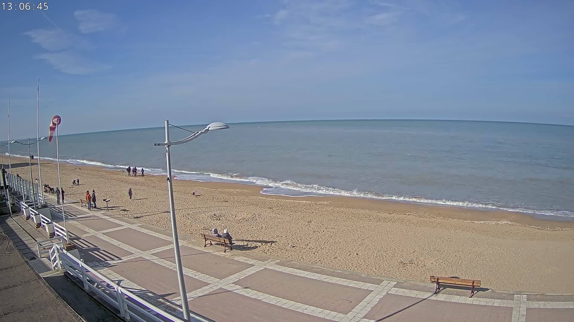 A sandy beach with a calm sea under a mostly clear sky is viewed from above, showing a paved walkway with several people strolling and sitting on benches.