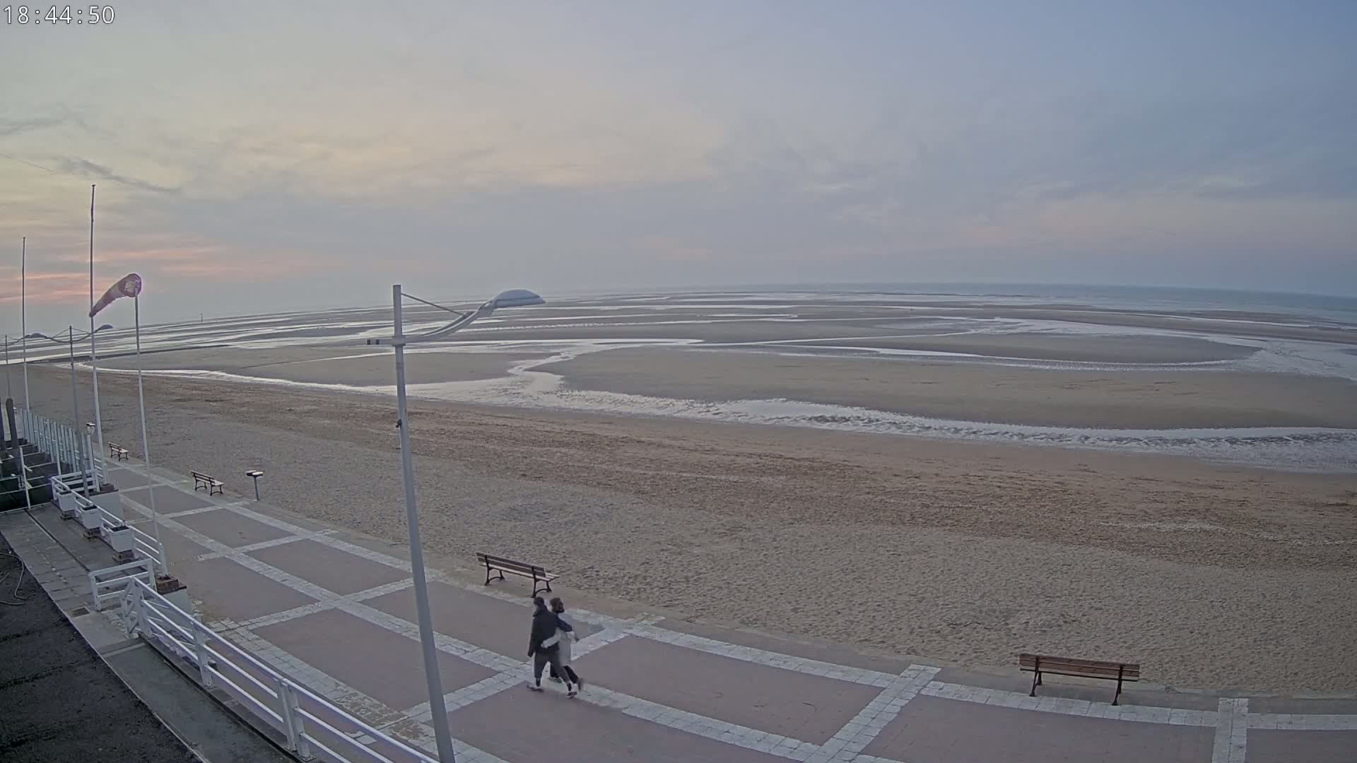 A couple walks along a paved beachfront path overlooking a wide, sandy beach at low tide under a pale, overcast sky.