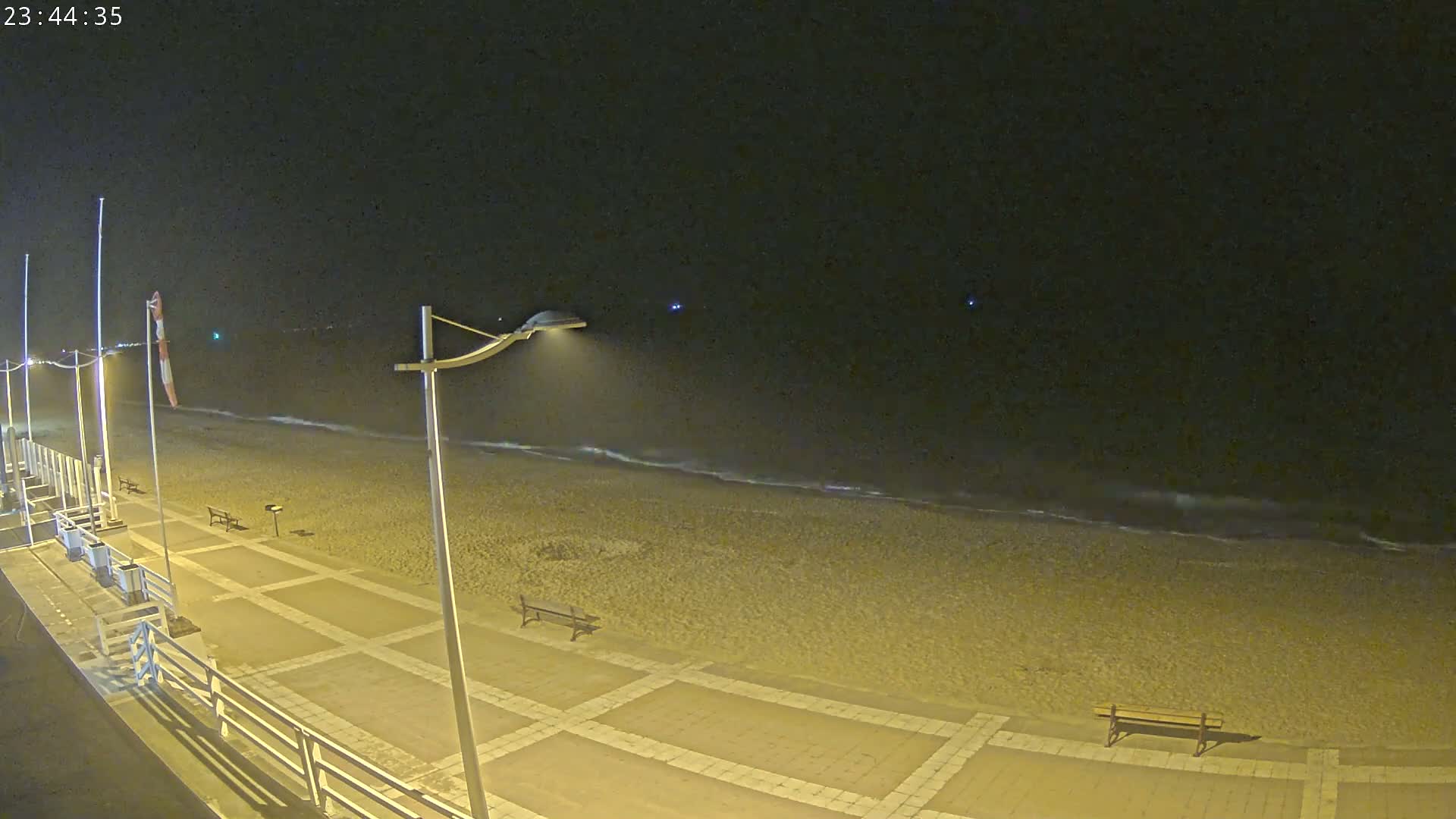 A nighttime view of a beach with a paved walkway, benches, and lampposts, under a dark sky with gentle waves.