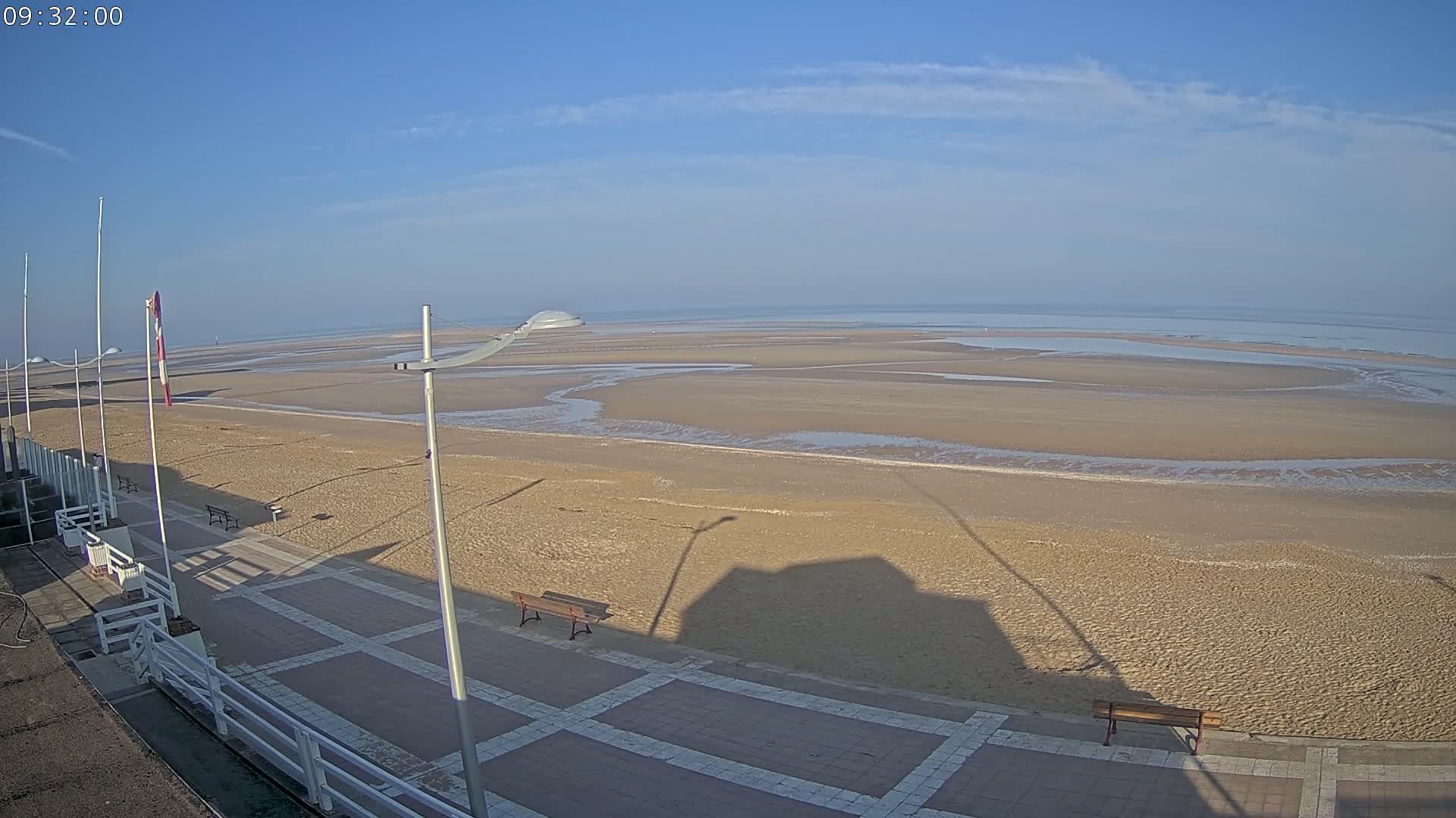A wide expanse of sandy beach at low tide stretches to a calm ocean under a clear, bright blue sky.