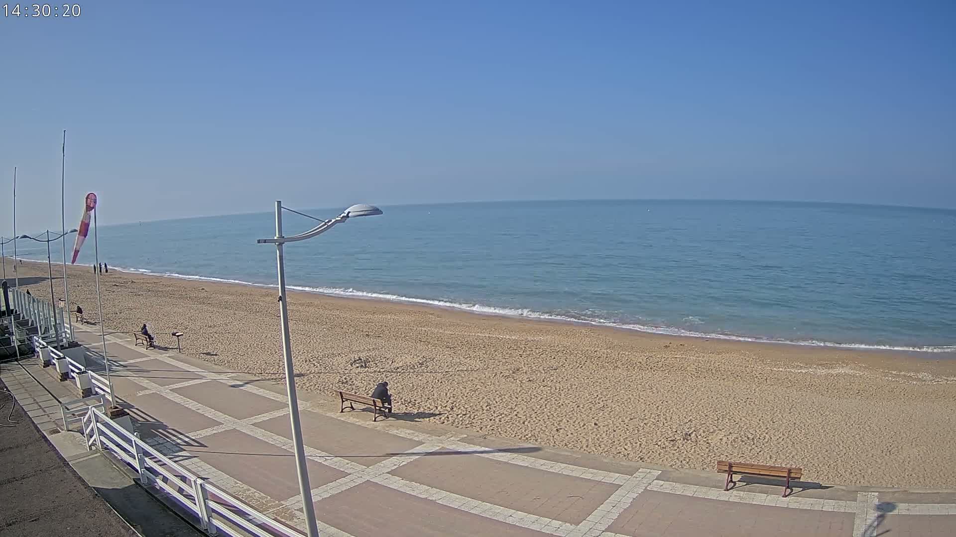 A sandy beach with calm blue water stretches out under a clear blue sky, and a paved walkway with benches runs along the shore.