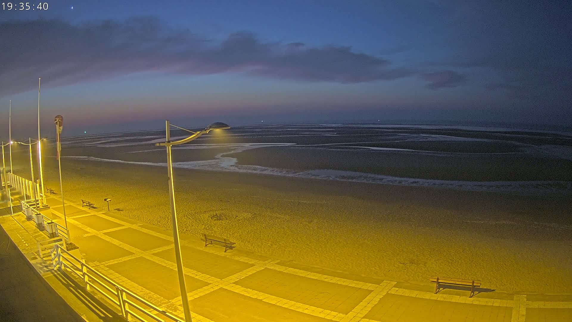 A nighttime view of a beach promenade with benches, illuminated by streetlights under a partly cloudy, dark blue sky.