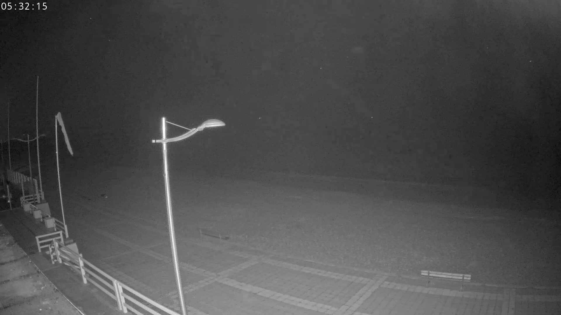 A nighttime, low-light view of an empty beach with a paved walkway, streetlights, and a bench visible.