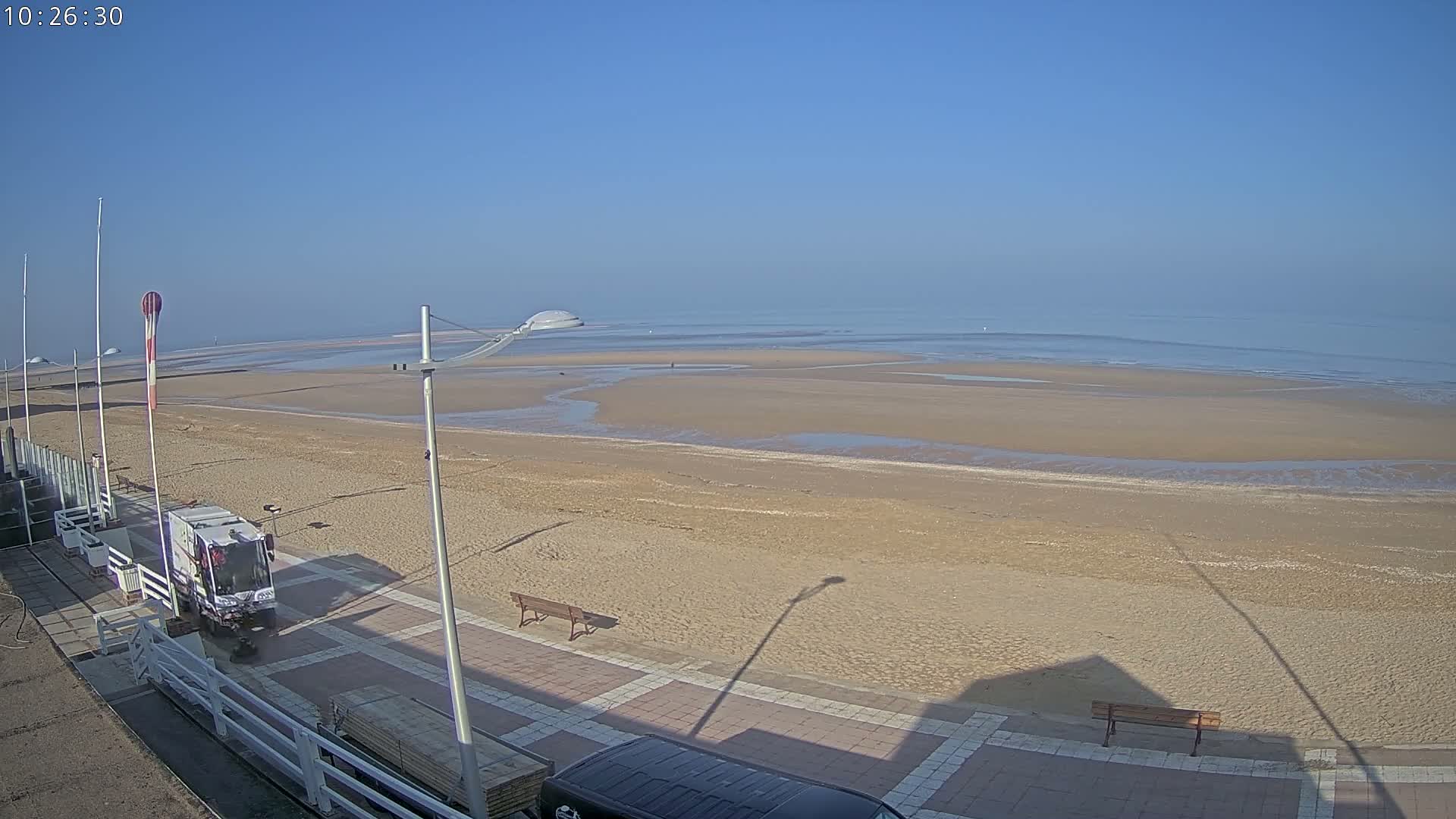 A beach with a mostly clear sky shows a wide expanse of sand and calm water, a beach cleaning vehicle, and a few park benches along a paved walkway.