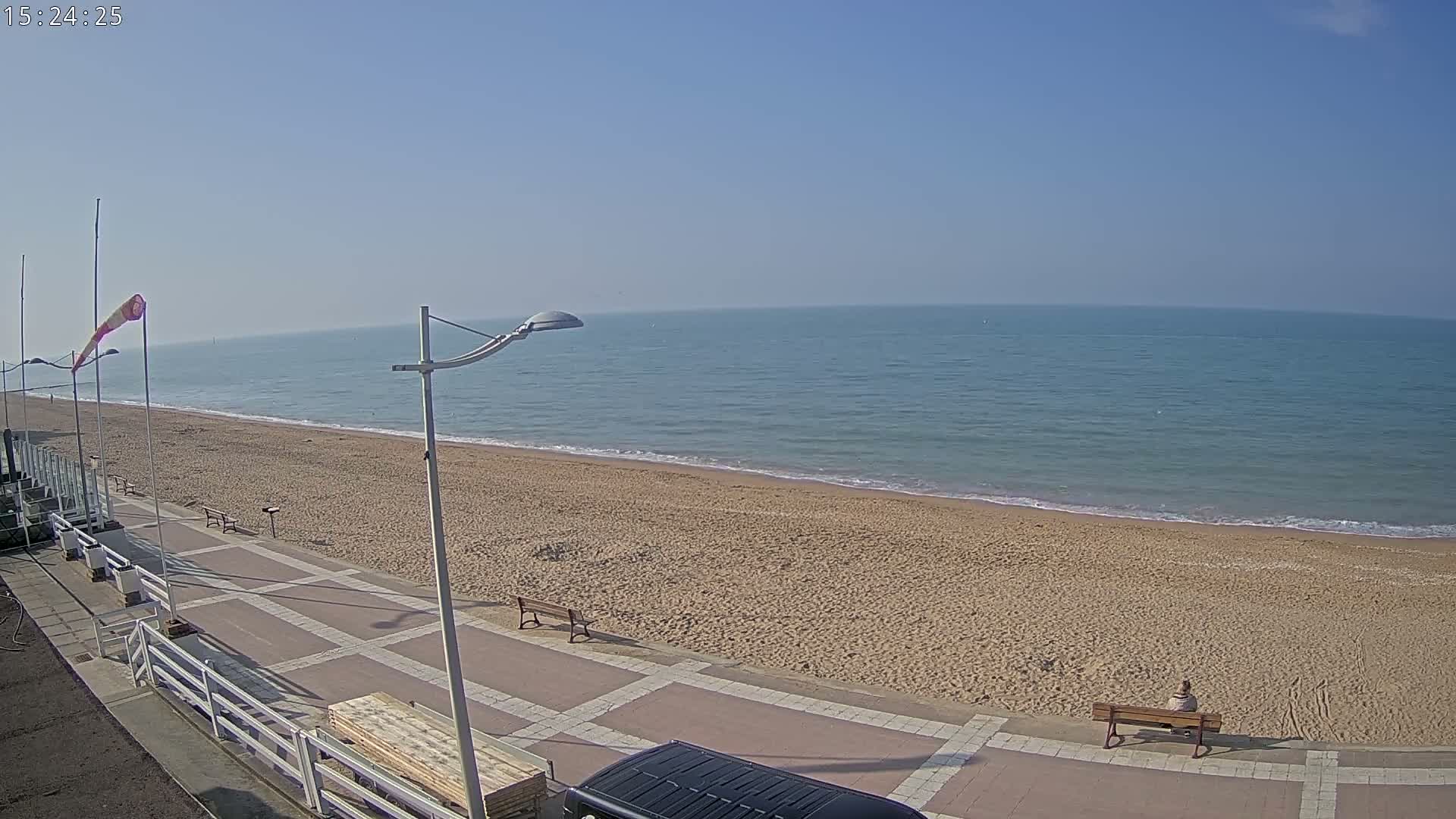 A sandy beach stretches along a calm ocean under a clear blue sky, with a paved walkway and benches nearby.