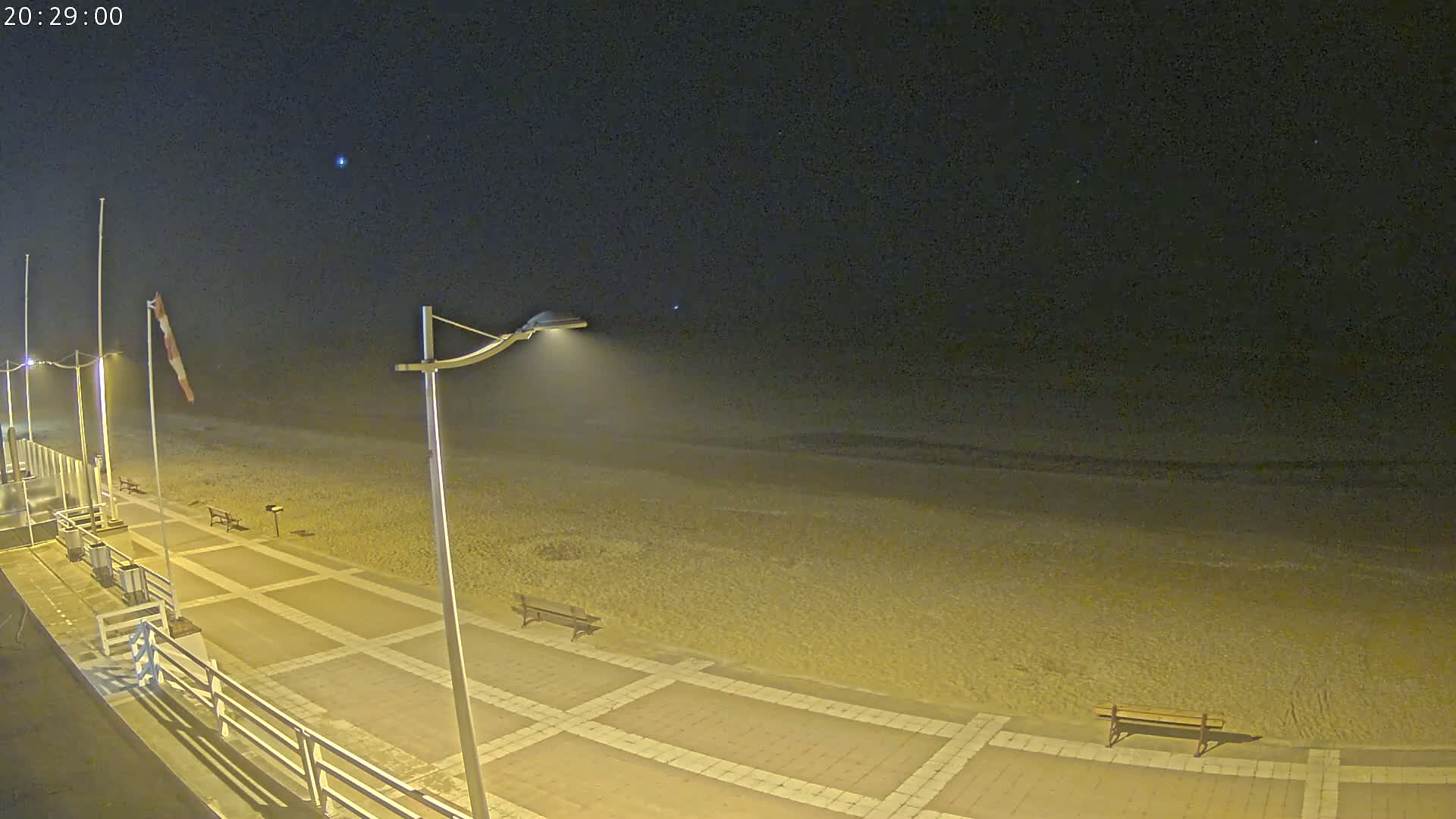 A nighttime view of a sandy beach with a paved walkway, streetlights, and benches under a clear, dark sky.