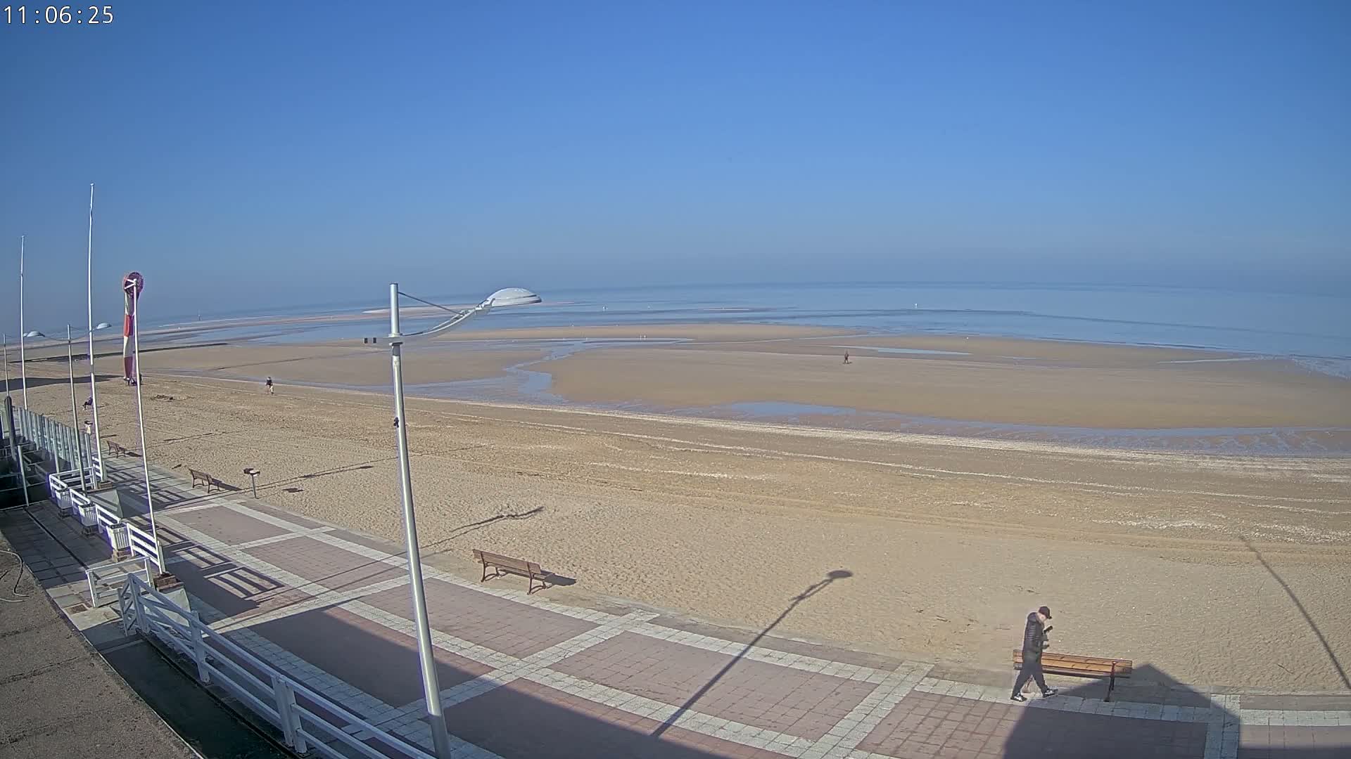 A mostly empty sandy beach stretches to a calm, light blue sea under a clear blue sky, with a paved walkway and a few people visible in the distance.