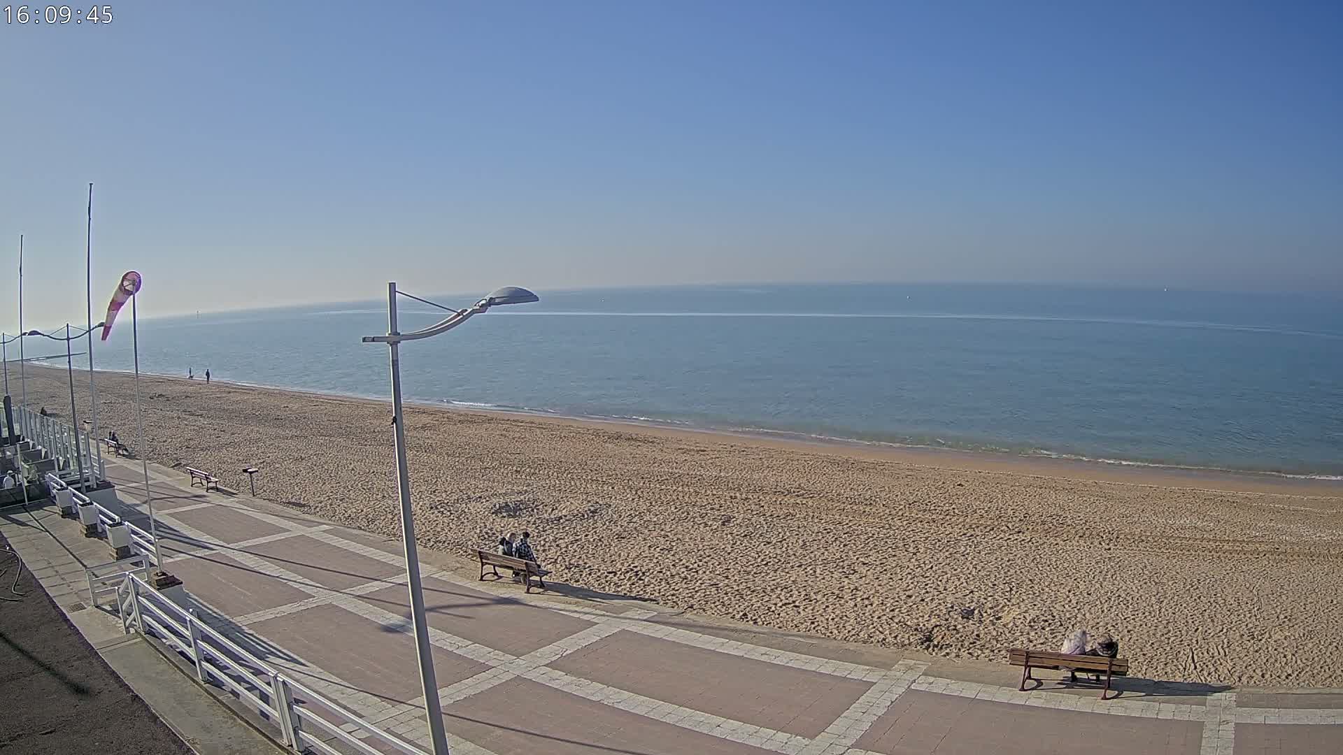 A mostly empty sandy beach stretches along a calm, blue ocean under a clear blue sky, with a paved walkway and a few people sitting on benches.