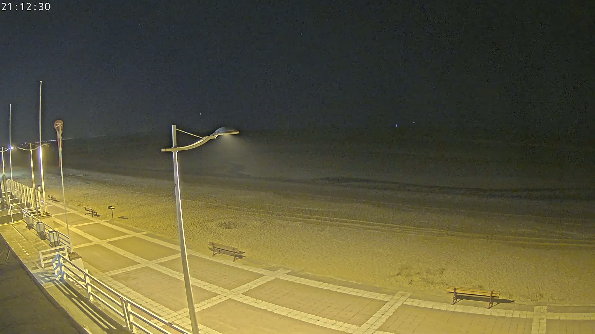 A nighttime view of a paved beachside walkway with benches, streetlights illuminating a sandy beach and calm ocean under a dark sky.