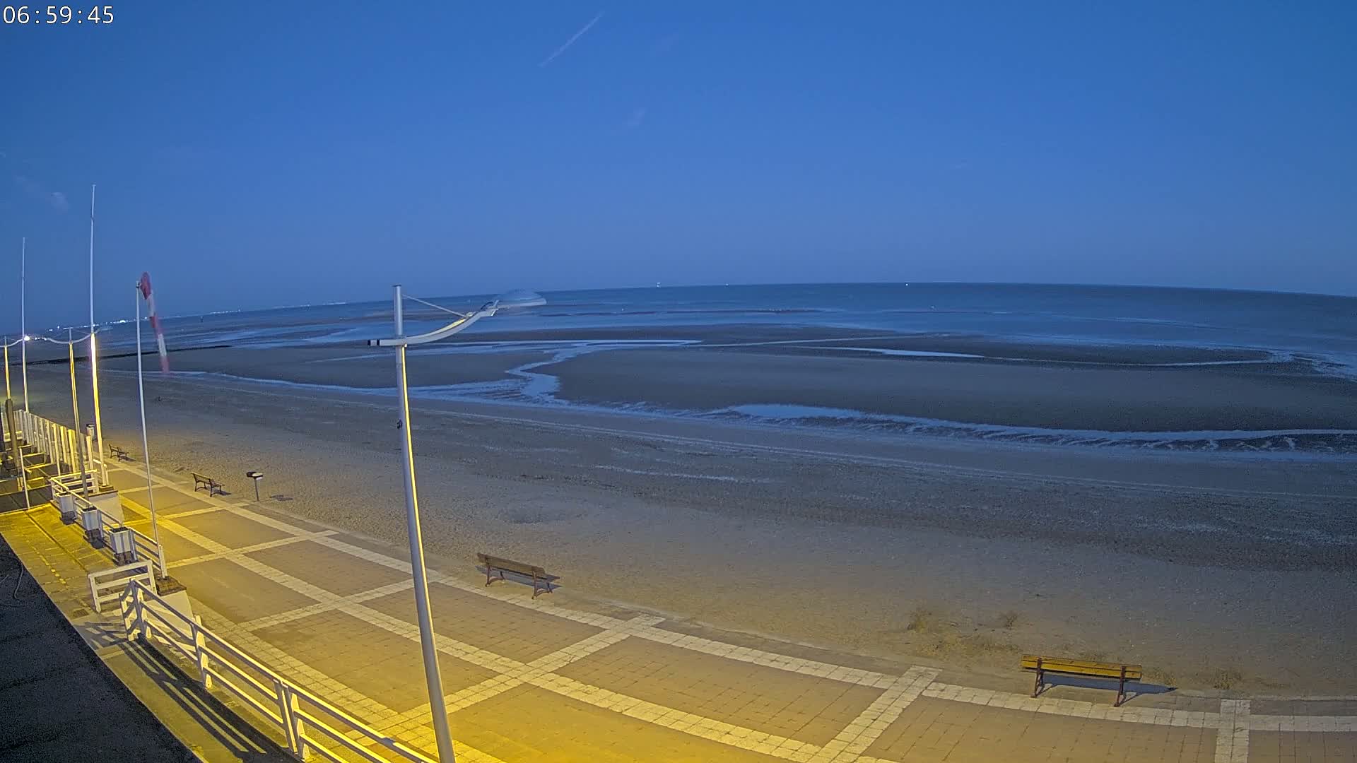 A mostly empty beach at twilight, with a paved walkway and benches, is illuminated by artificial light.