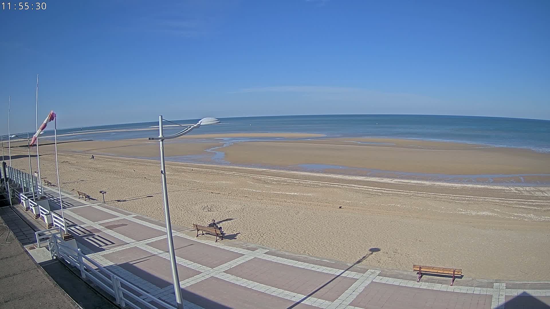 A mostly sunny day at a beach shows a wide sandy expanse leading to calm ocean water; a paved walkway with benches runs along the beach.