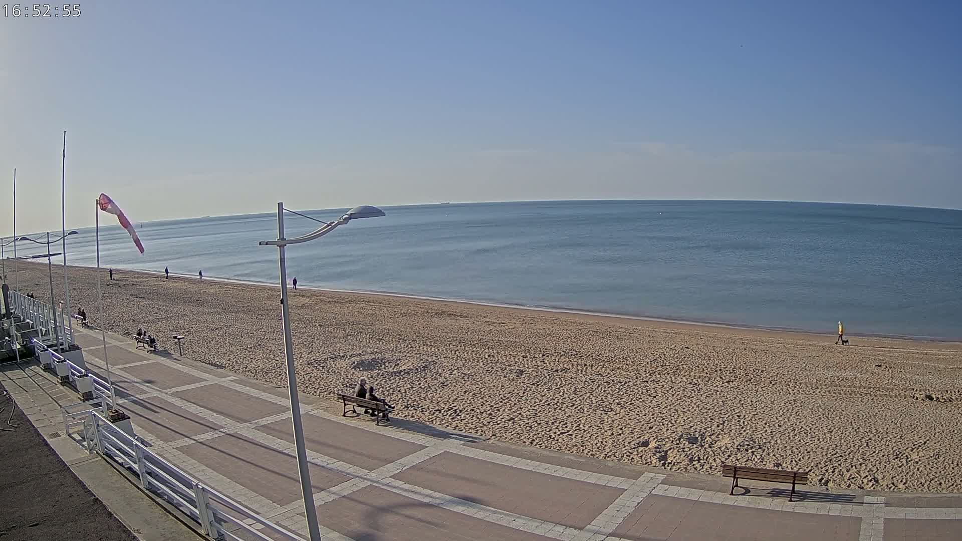 A mostly sunny day at a beach shows a wide sandy shore, calm ocean water, and a paved walkway with benches and a few people.