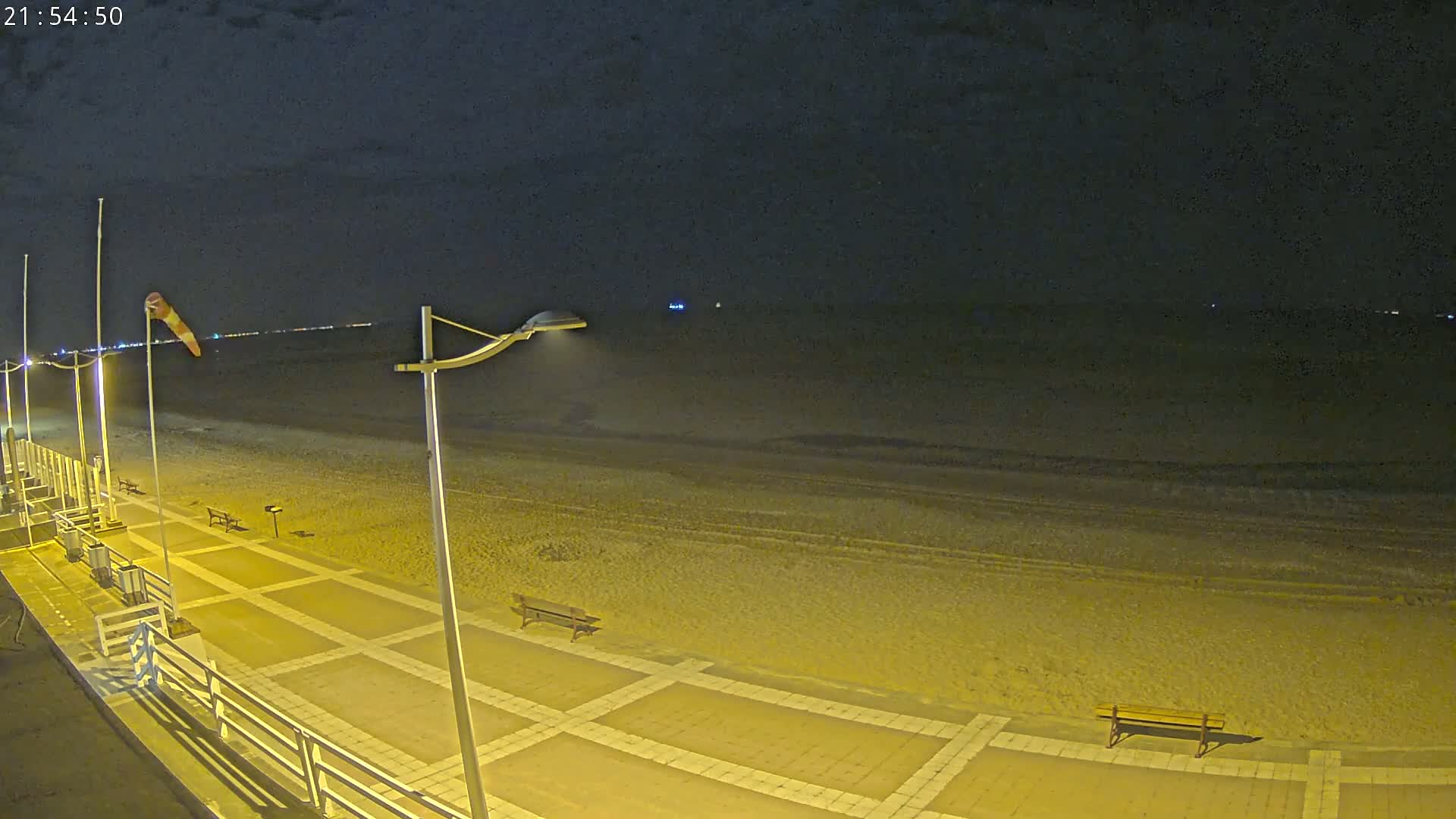 A nighttime view of a mostly empty beach boardwalk with a windsock and several lights, overlooking a dark ocean under a clear sky.