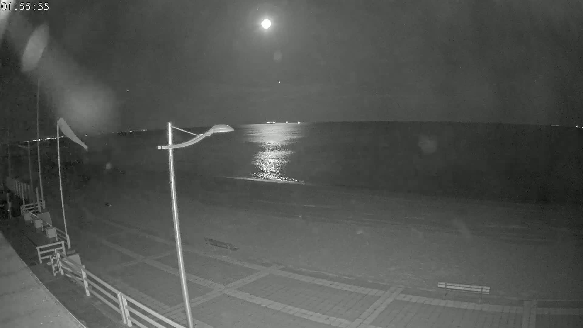 A moonlit beach at night, showing a calm sea reflecting the light, with a paved walkway and a lamppost in the foreground.