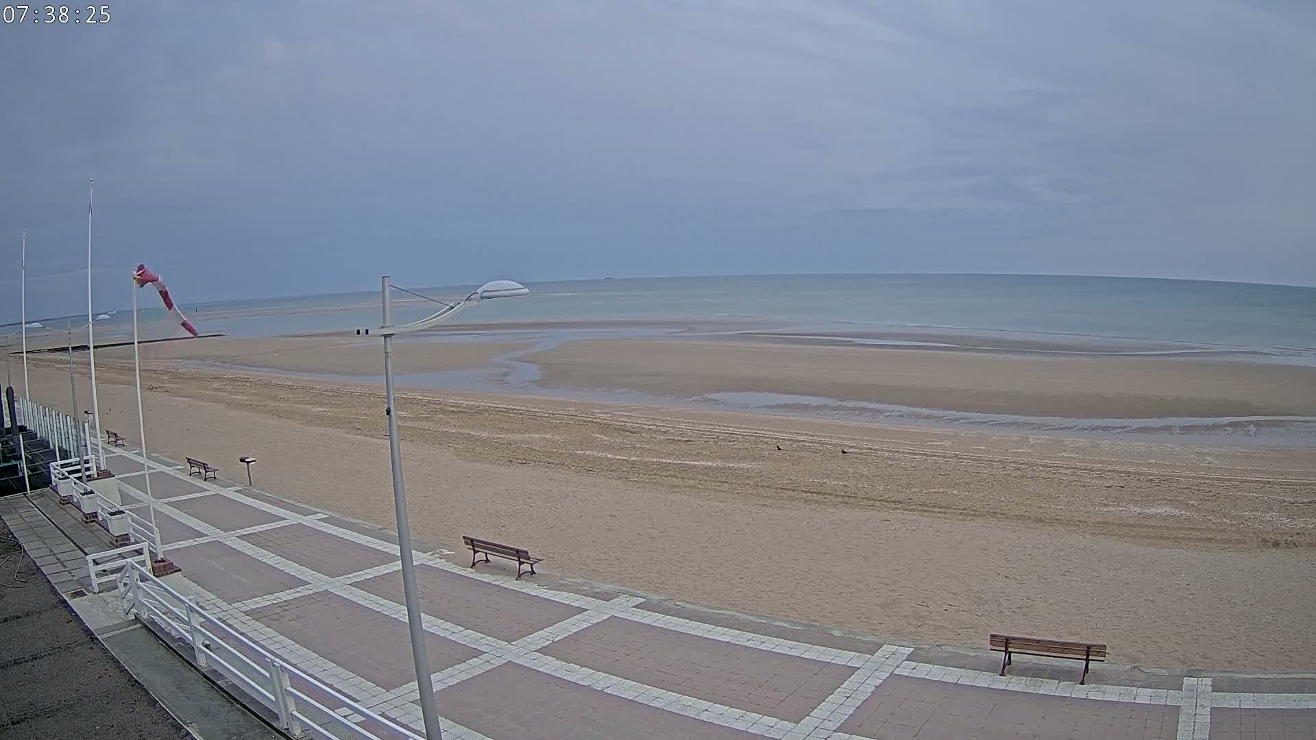A mostly sunny day at a beach shows a wide sandy shore receding into a calm sea, with a paved walkway and benches visible in the foreground.