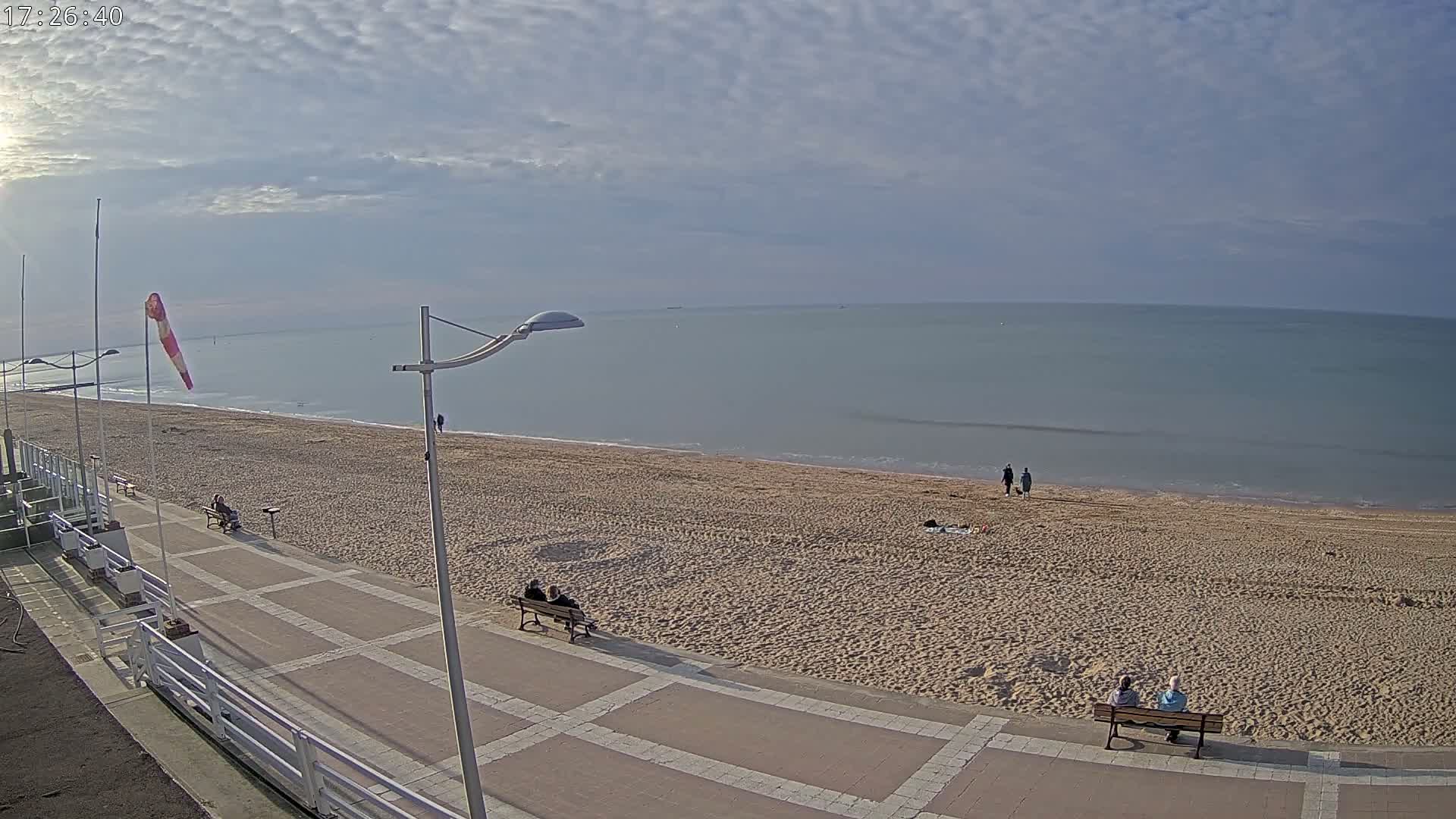 A mostly sunny day shows a sandy beach with a few people sitting on benches and walking along a paved walkway next to a calm ocean.