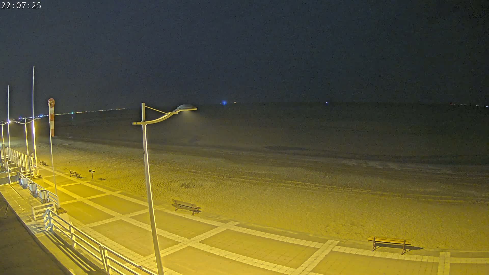 A nighttime view of a beach with a paved walkway, benches, and lampposts, under a dark sky.