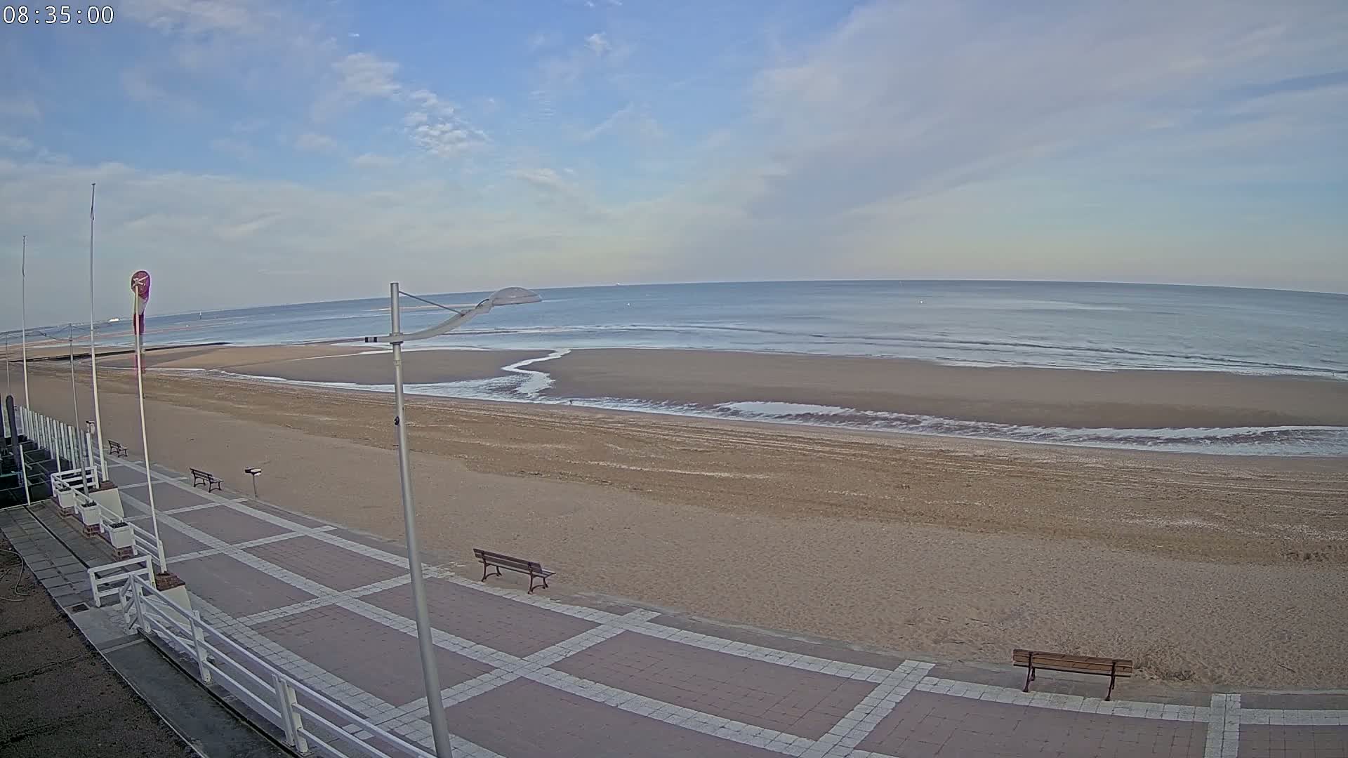 A mostly sunny day shows a wide sandy beach with small waves lapping the shore, and a paved walkway with benches leading from a beachfront promenade.