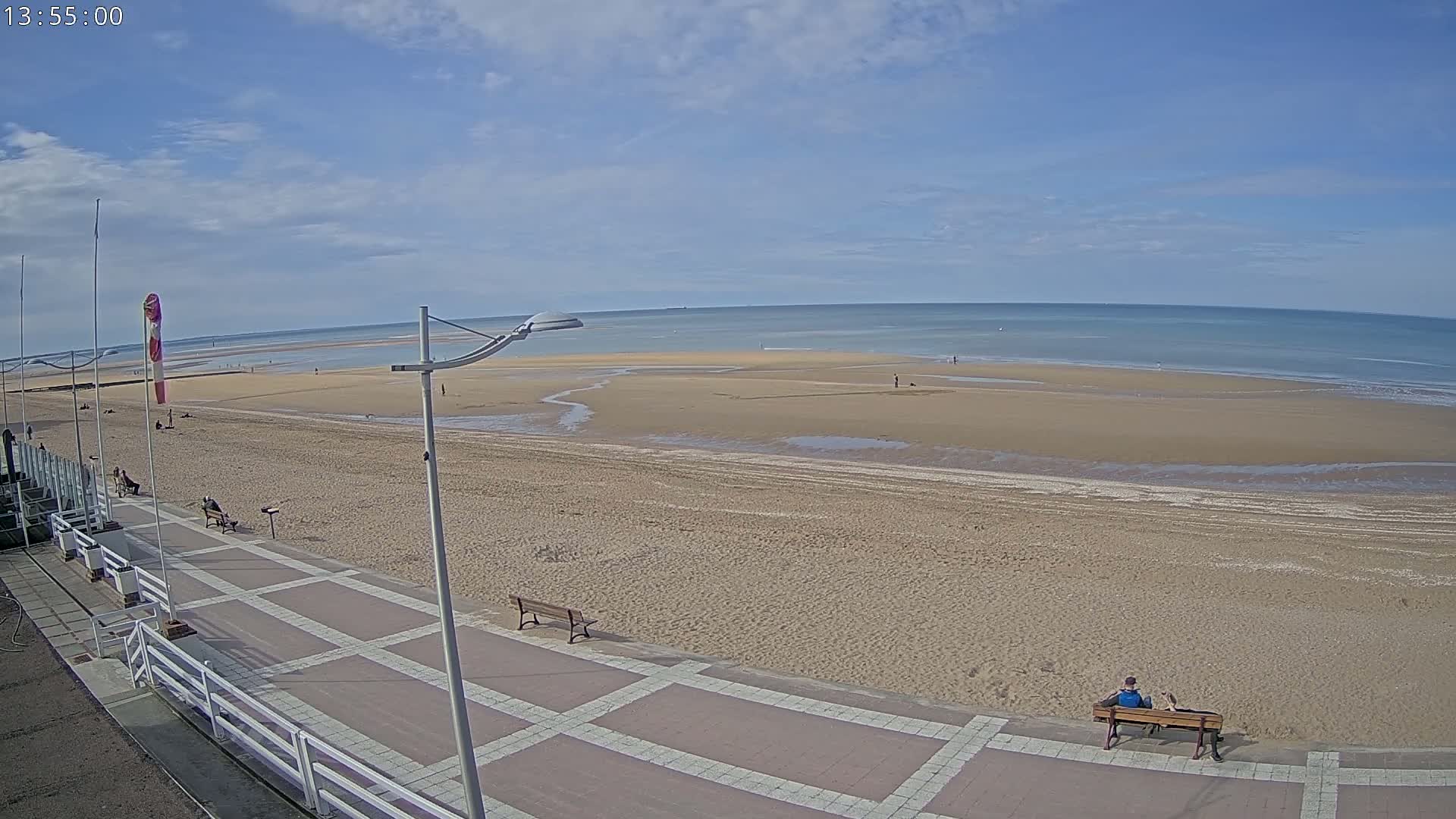 A sandy beach with a paved walkway and a few people, under a partly cloudy sky, stretches to a calm ocean.