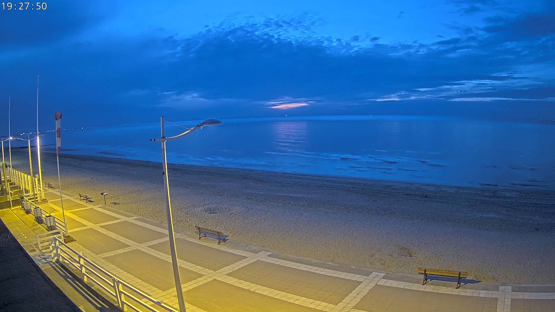 A nighttime beach scene under a partly cloudy, dark blue sky shows a paved walkway with benches overlooking a calm, dark-blue ocean.