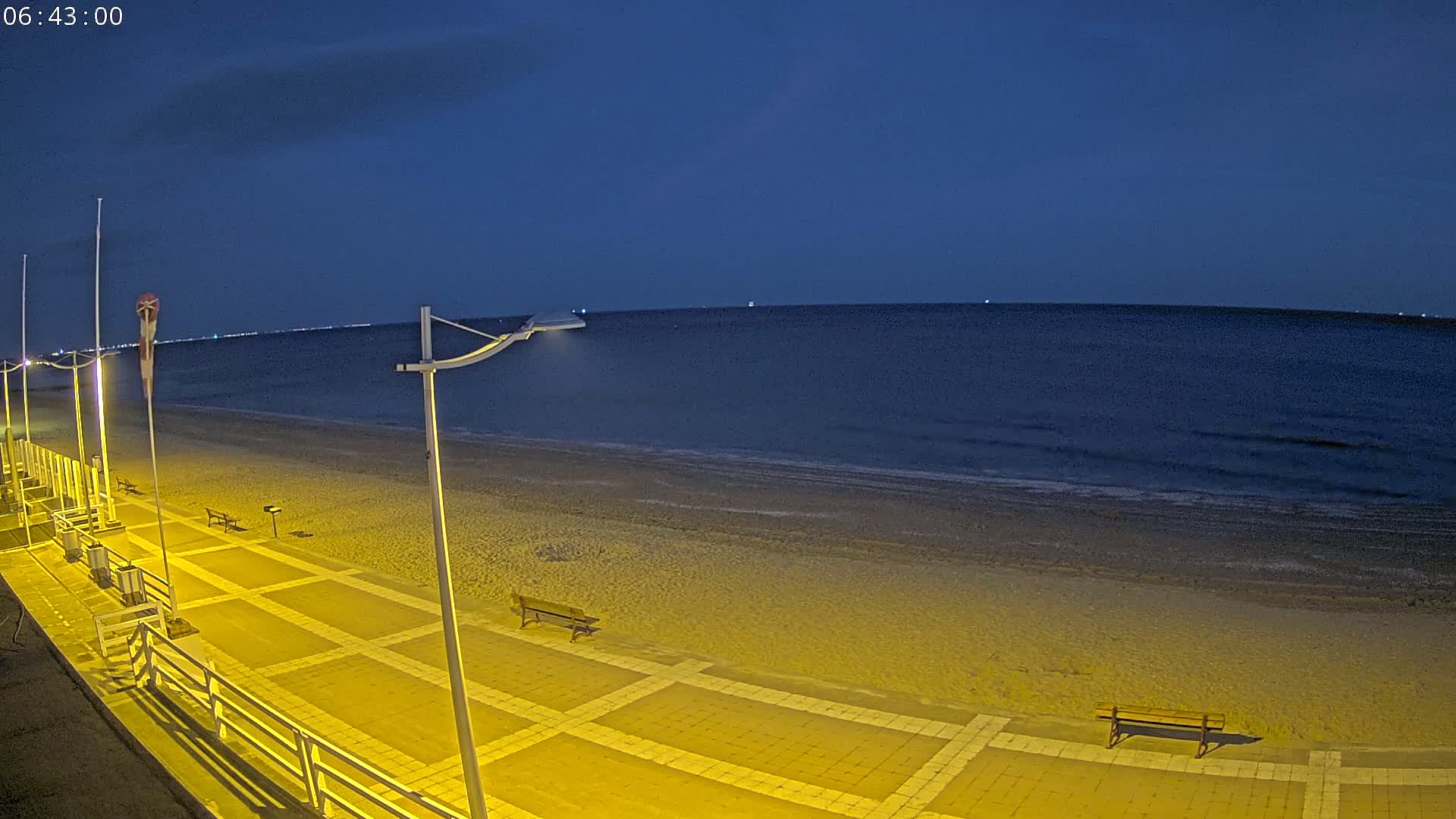 A sandy beach at night under a dark blue sky is illuminated by yellow lights along a paved walkway with benches.