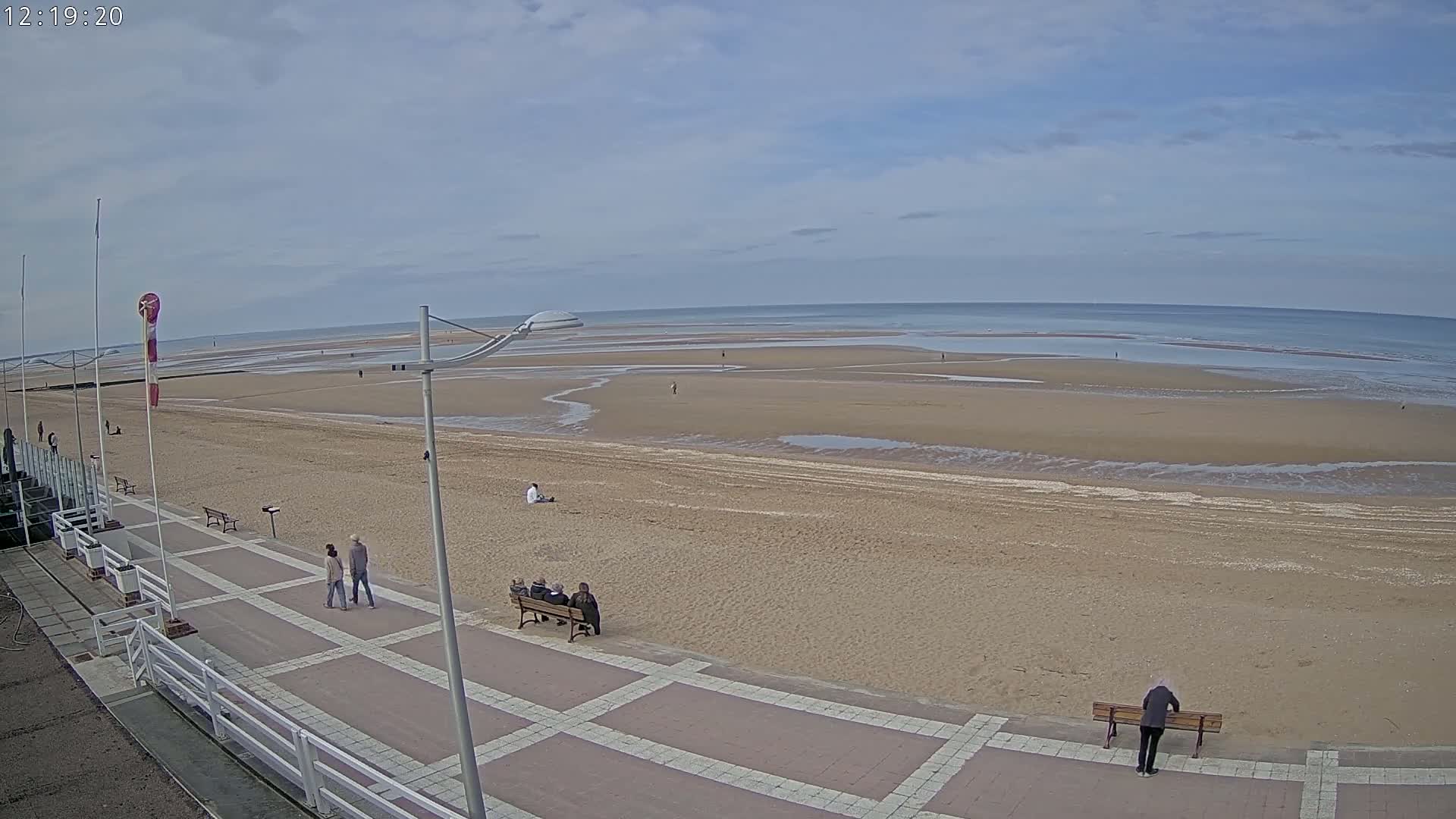 A wide sandy beach stretches to a calm ocean under a mostly cloudy sky, with a paved walkway and several people visible.