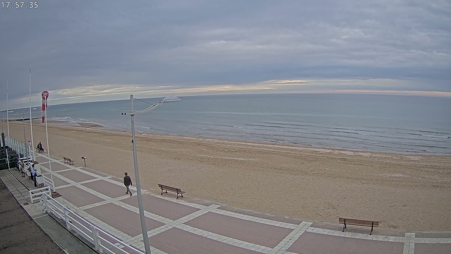 A sandy beach with a paved walkway and several people walking, under a cloudy sky with a calm sea in the background.