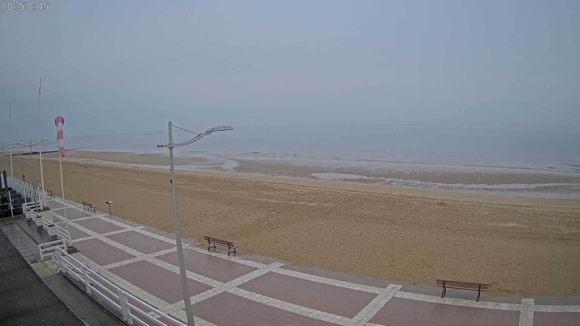 A mostly empty sandy beach with a paved walkway and benches is visible on an overcast day.