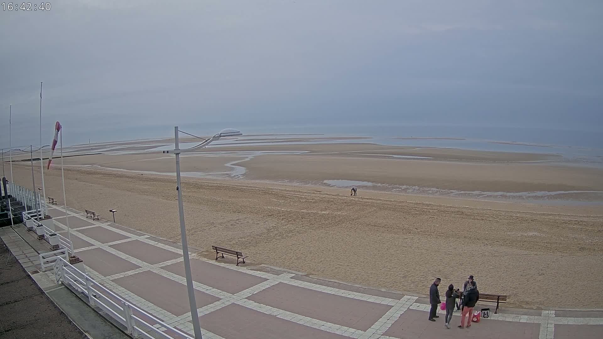 A wide sandy beach stretches to a calm, gray sea under an overcast sky, with a paved walkway and several people near a bench in the foreground.