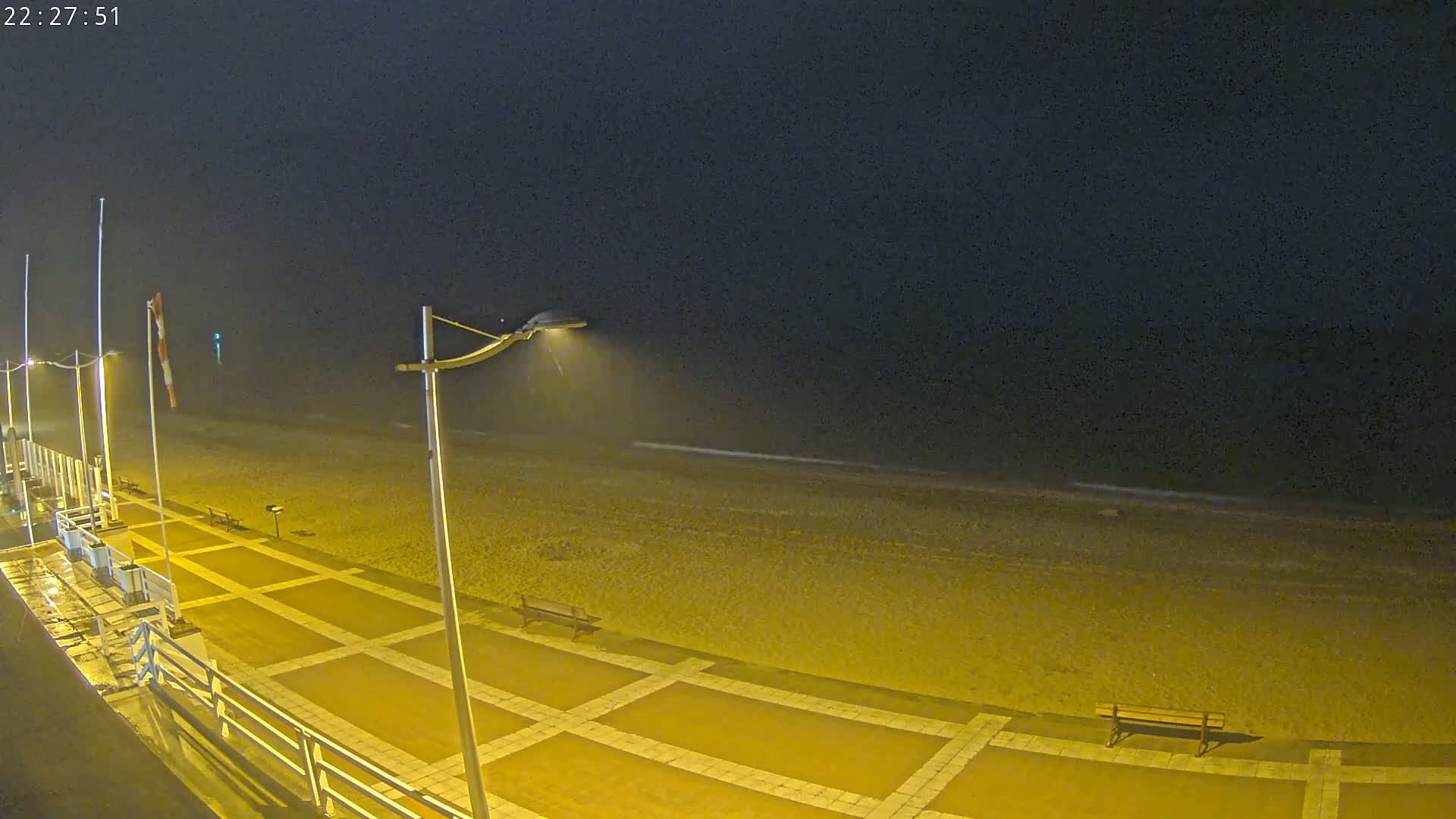 A nighttime view of a mostly empty beach with a paved walkway, lampposts, and a bench, under a dark, possibly misty sky.