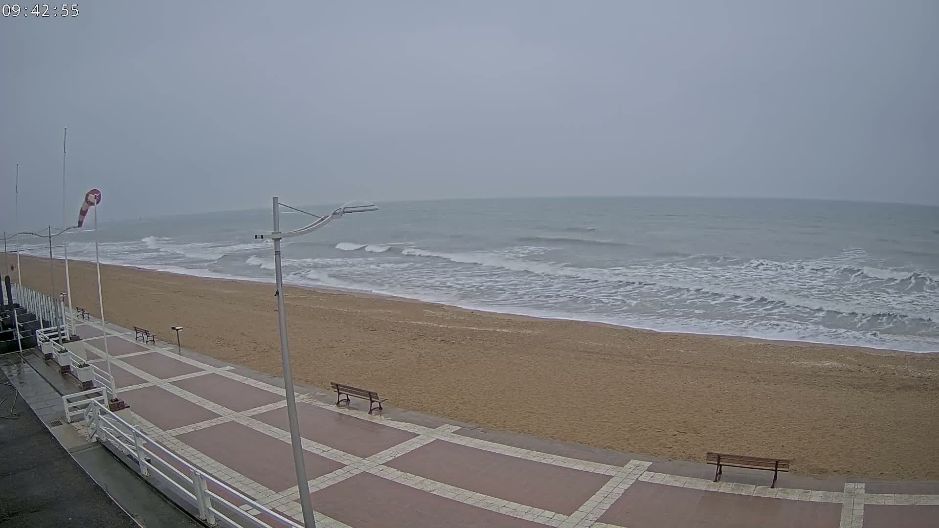 A sandy beach with moderate waves under an overcast sky is viewed from a paved walkway with benches.