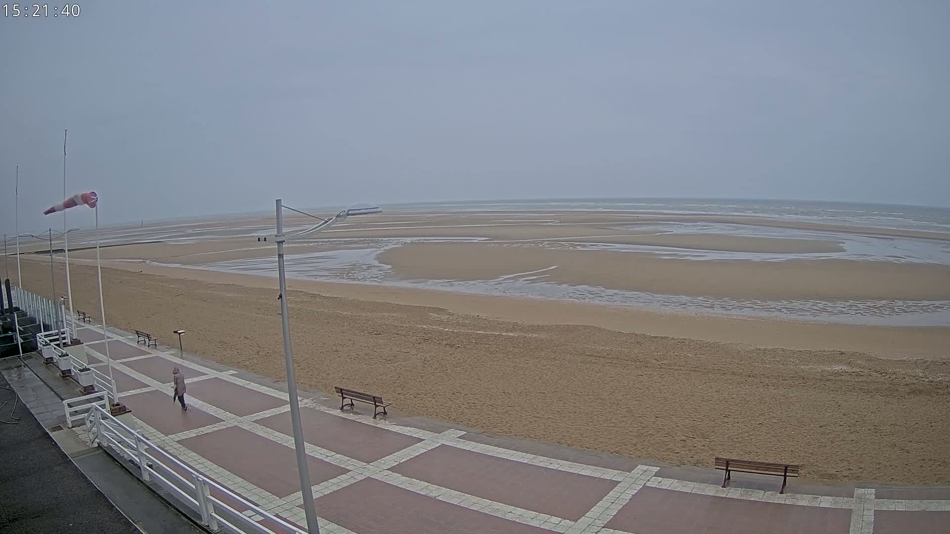 A person walks along a paved beachfront walkway overlooking a wide, sandy beach with shallow, receding water under an overcast sky.
