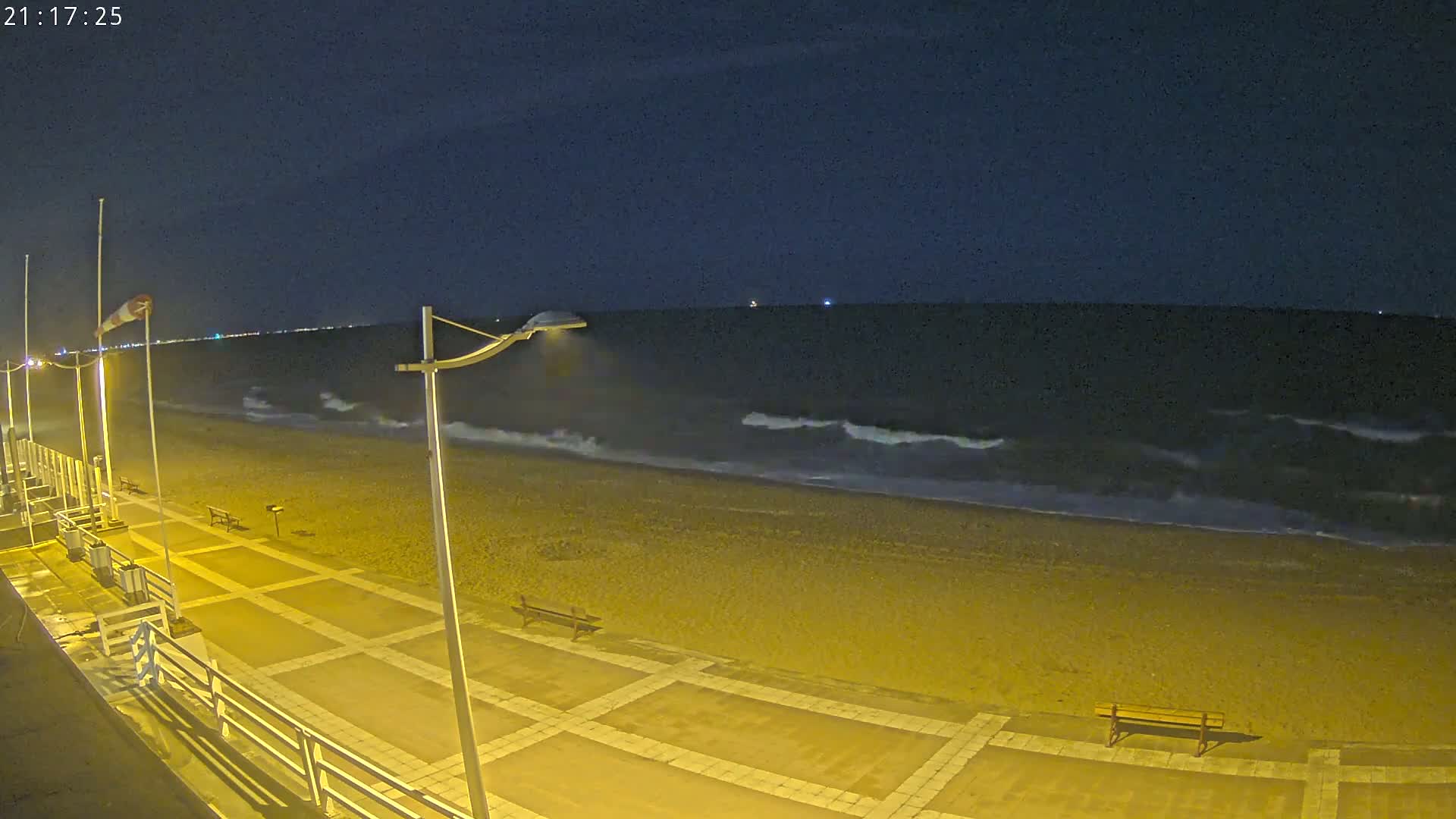 At night, under a dark sky, moderately sized waves crash on a pebble beach next to a paved walkway with benches and streetlights.