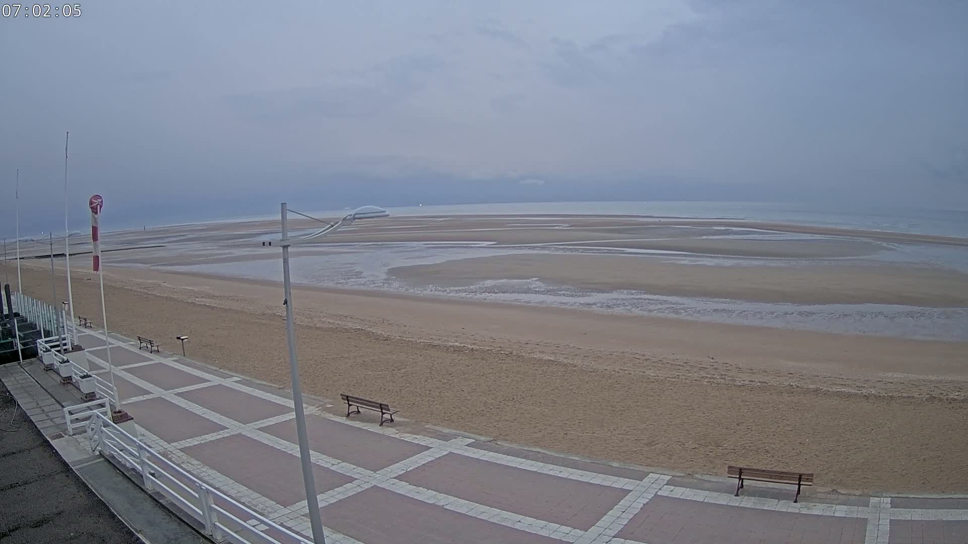 A wide sandy beach stretches to a calm, overcast ocean, with a paved walkway and benches visible in the foreground.