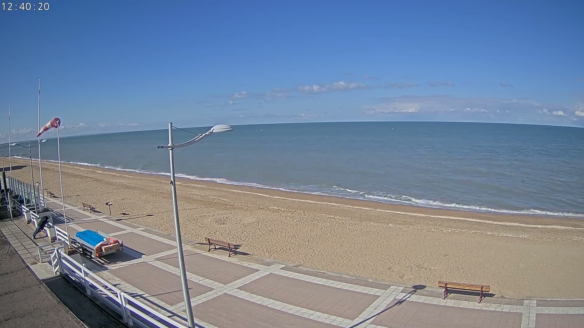 A sandy beach with a paved walkway and benches stretches along a calm sea under a mostly clear blue sky.