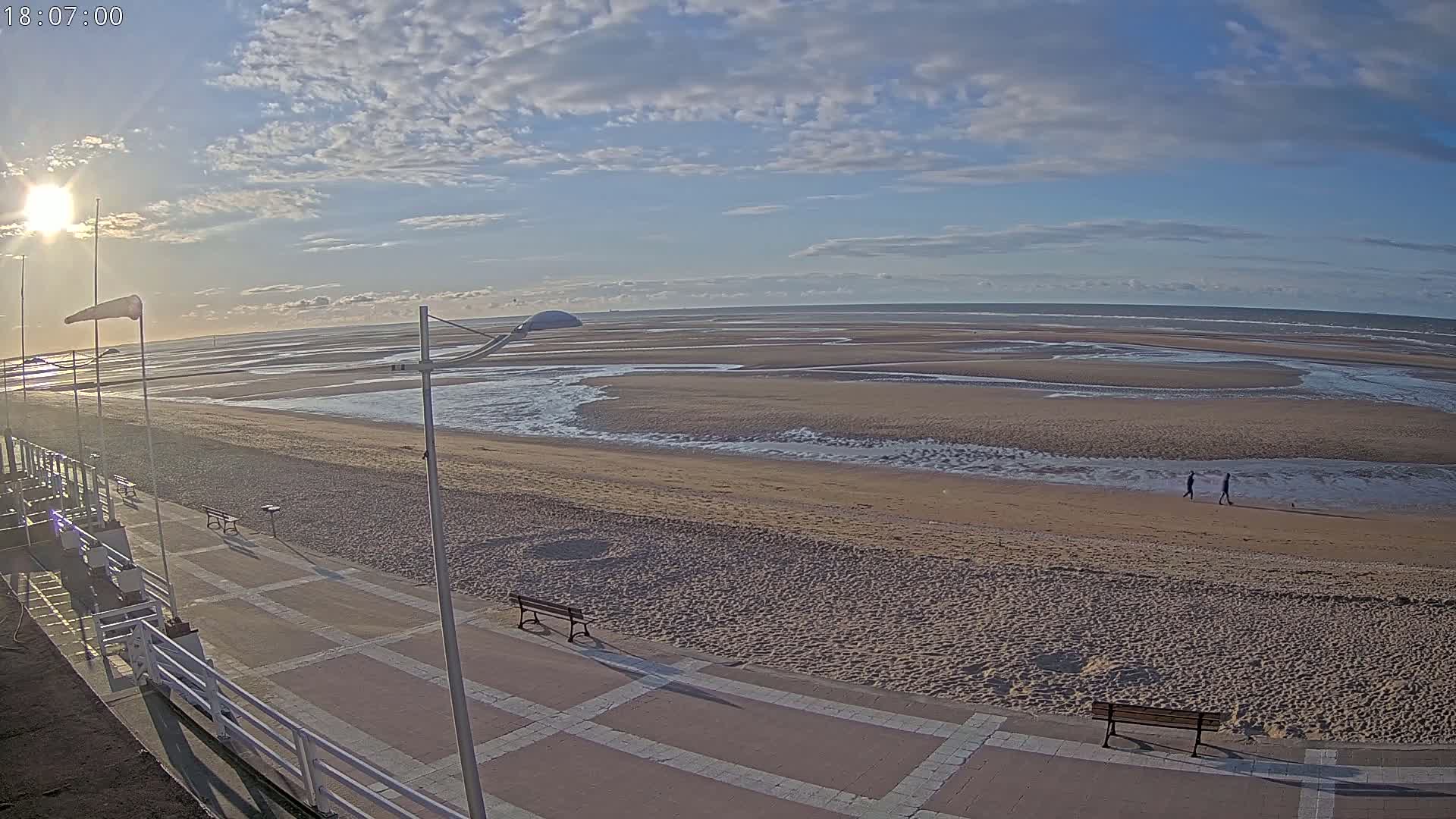 A wide sandy beach stretches to a calm ocean under a partly cloudy sky, with a paved boardwalk and benches in the foreground.