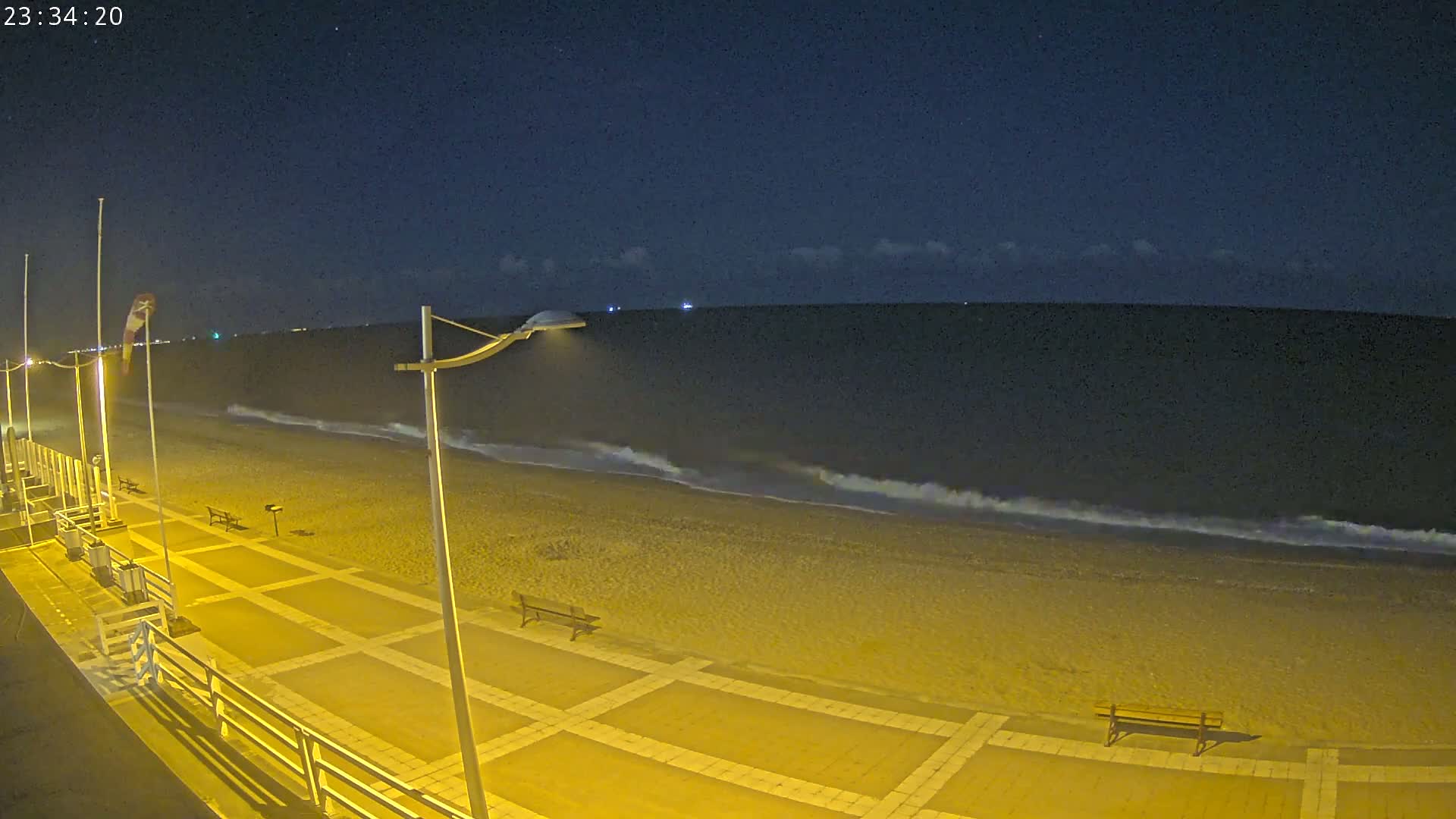 A nighttime beach scene shows a paved walkway next to a sandy beach with gentle waves under a mostly clear, dark sky.