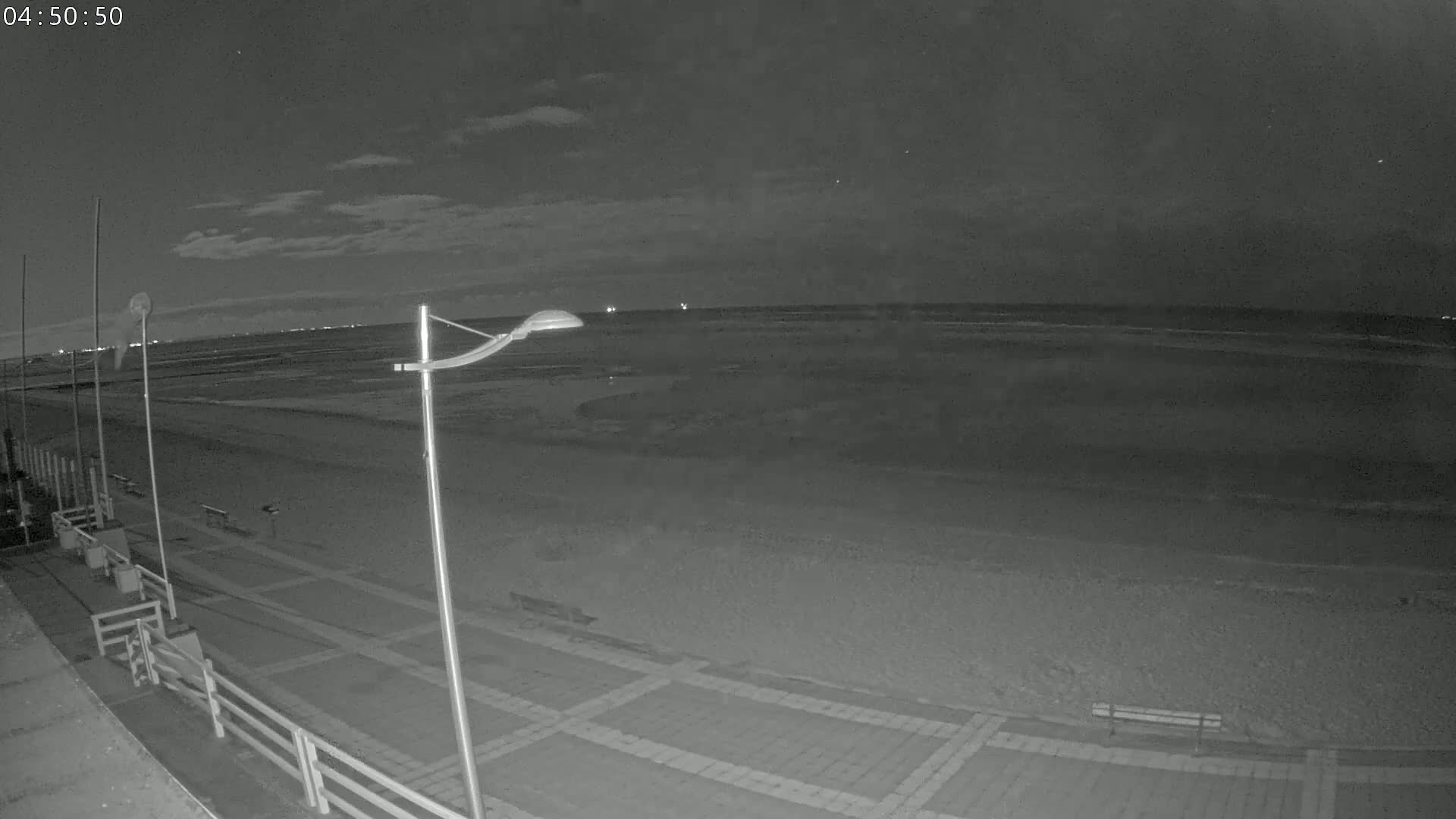 A nighttime view of a mostly empty beach with a paved walkway and lampposts, under a partly cloudy sky.