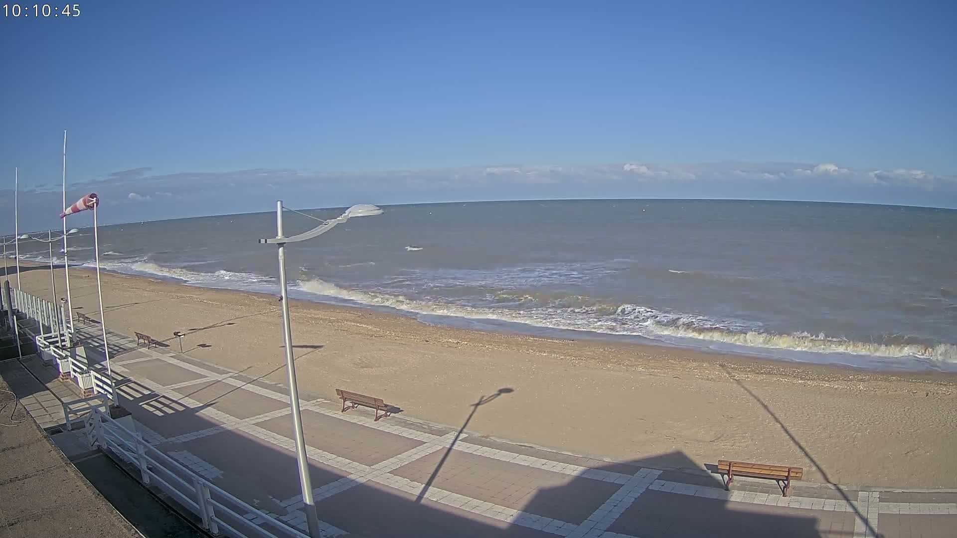 A mostly sunny day shows a sandy beach with gentle waves lapping the shore, a paved walkway with benches, and a windsock indicating wind.