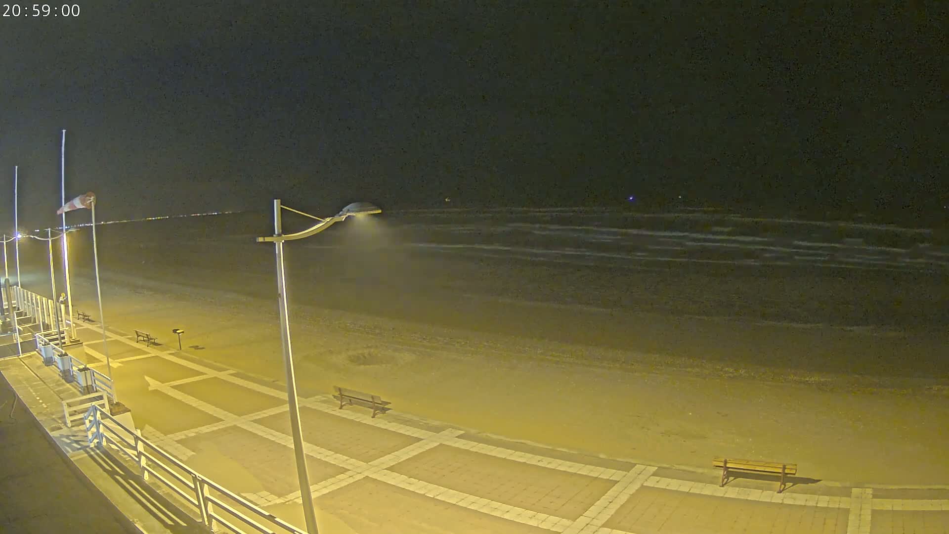 A nighttime view of a sandy beach with gentle waves under a dark sky, showing a paved walkway with benches and streetlights along a boardwalk.